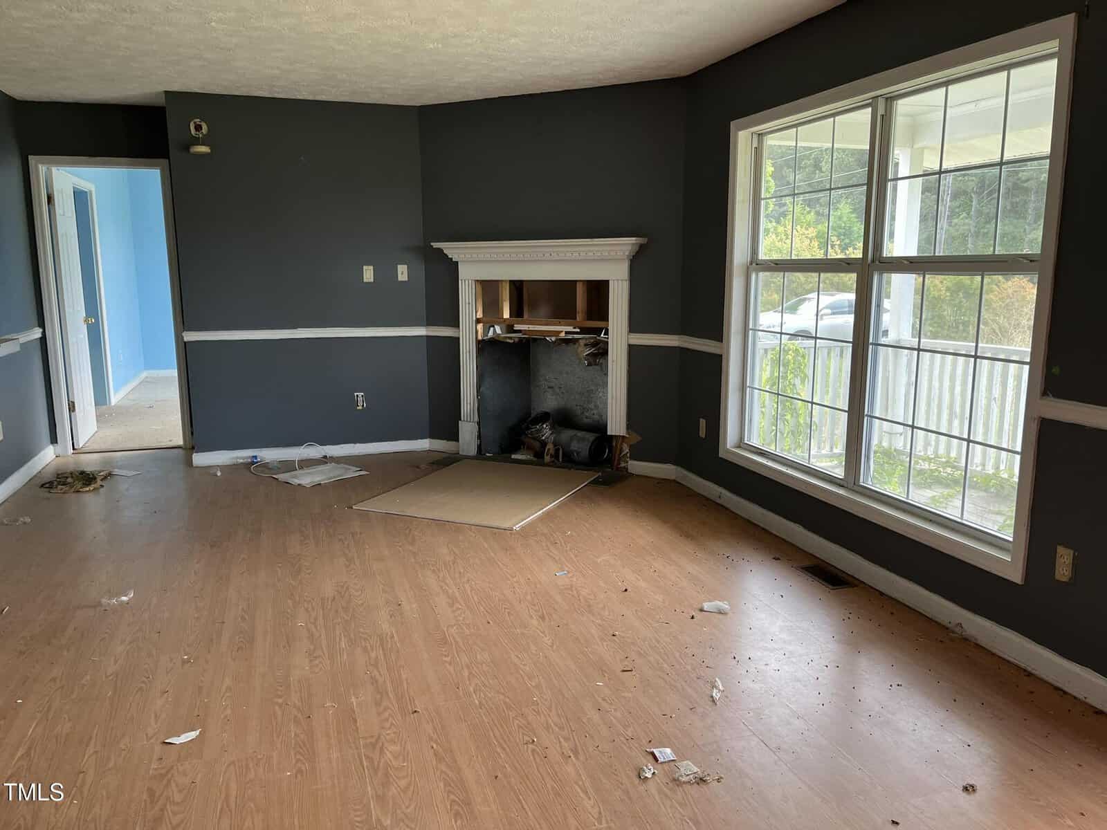 A mostly empty room with dark gray walls, light wood flooring, a fireplace missing its cover, scattered debris on the floor, a large window, and an open doorway leading to a blue-painted room.