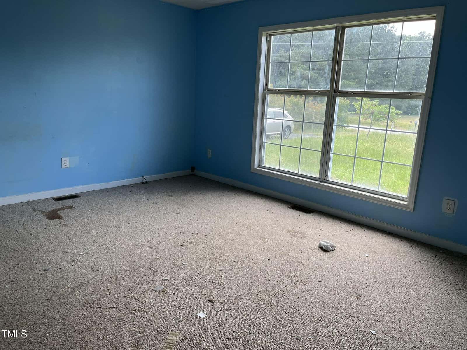 An empty room with light blue walls, beige carpet with stains and debris, two visible floor vents, and a large window showing a grassy yard and a parked car outside.