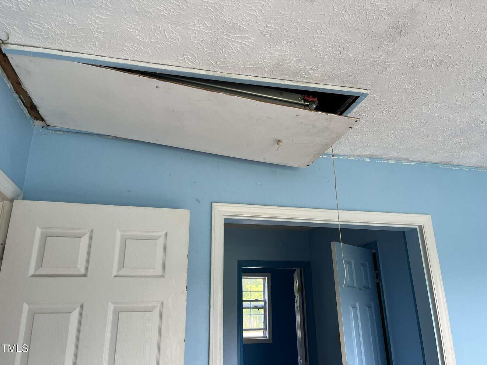 A white attic access panel in the ceiling is partially open, with a pull cord hanging down. The room has blue walls, white trim, and two doors; one door is open, revealing another blue room beyond.