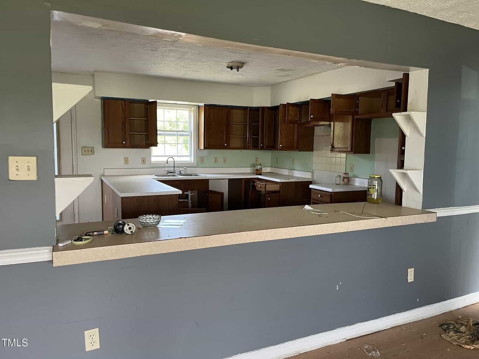 A view of an empty kitchen with open wooden cabinets, a countertop bar, a sink beneath a window, and shelves on each side of the pass-through. The kitchen appears dated and in need of cleaning or renovation.