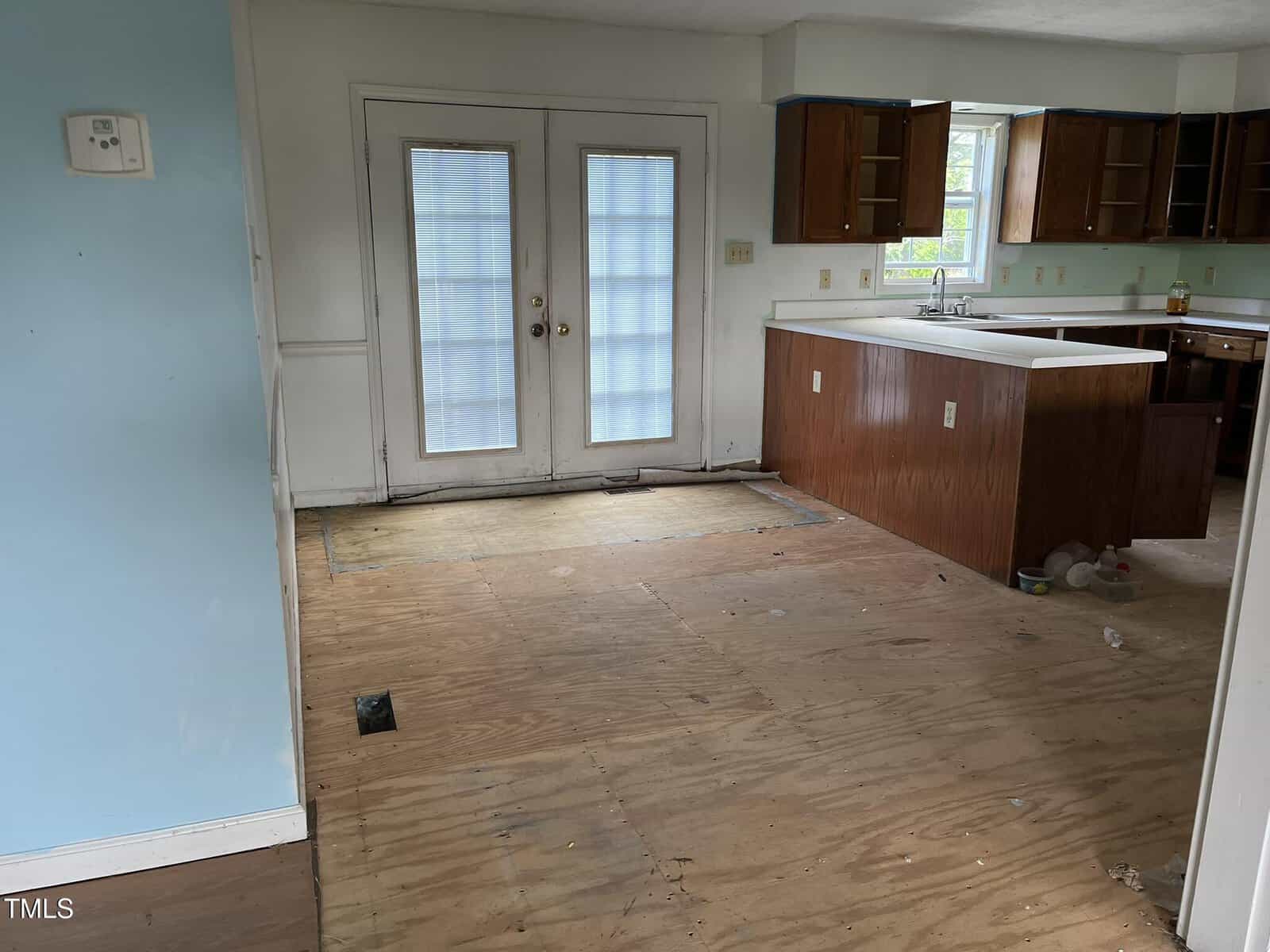A room with unfinished plywood flooring, empty lower shelves, and upper kitchen cabinets. French doors with blinds lead outside. Some debris is scattered on the floor near the kitchen area.