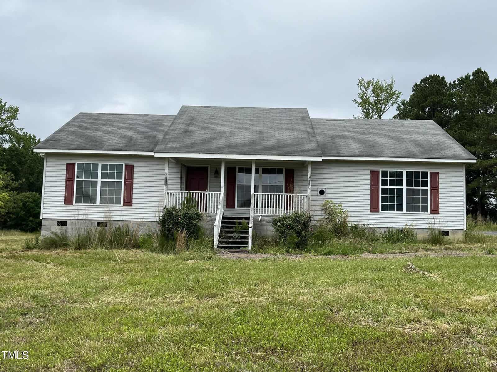 A single-story white house at 3213 Sam Usry Road, Oxford, features a front porch, two red doors, and four steps leading up. The yard is overgrown with tall grass beneath an overcast sky, with bushes near the front windows.
