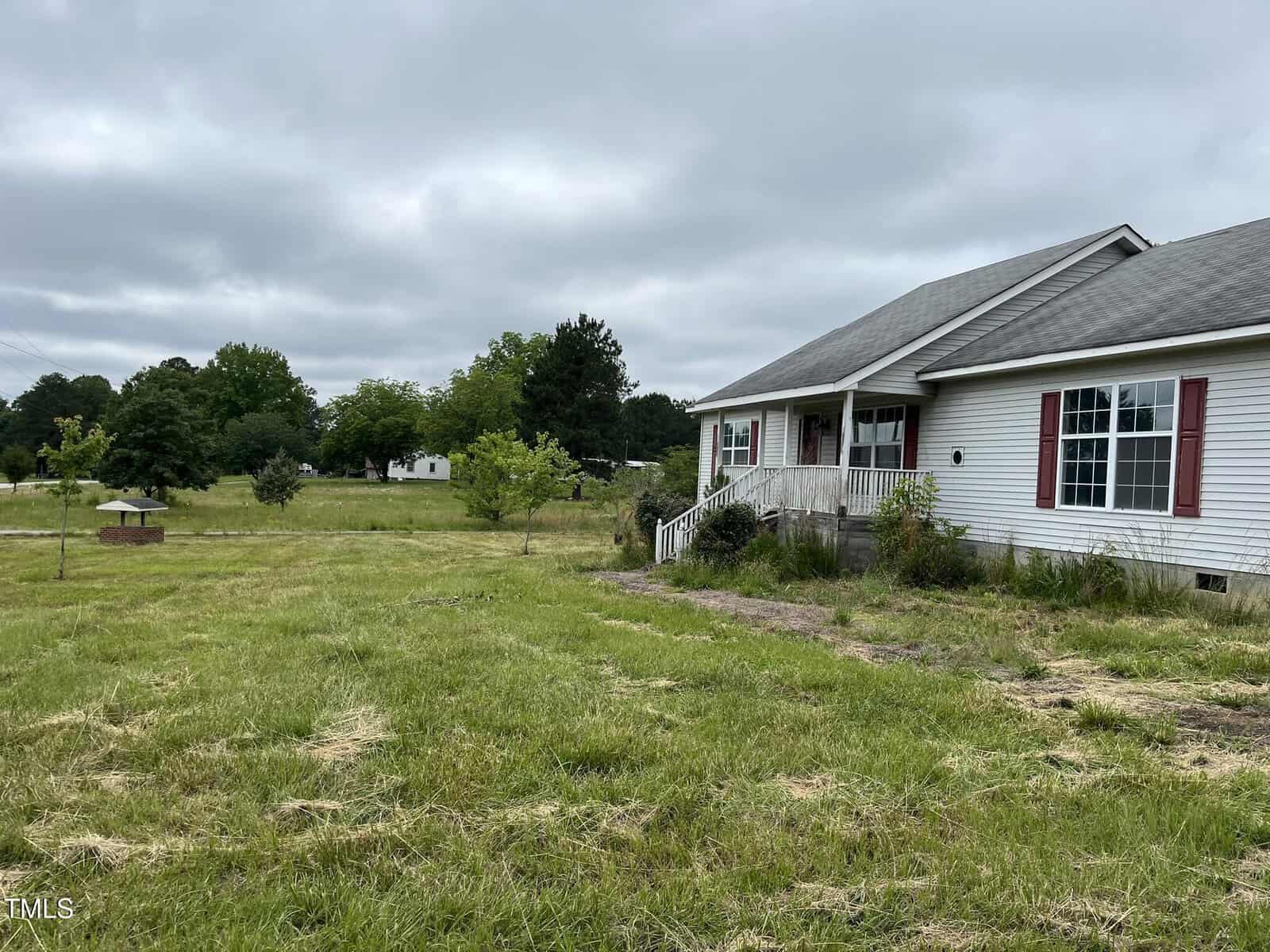 A white single-story house with red shutters sits on a large grassy lot with a few small trees. There is a covered well in the yard and another house visible in the background. The sky is overcast.
