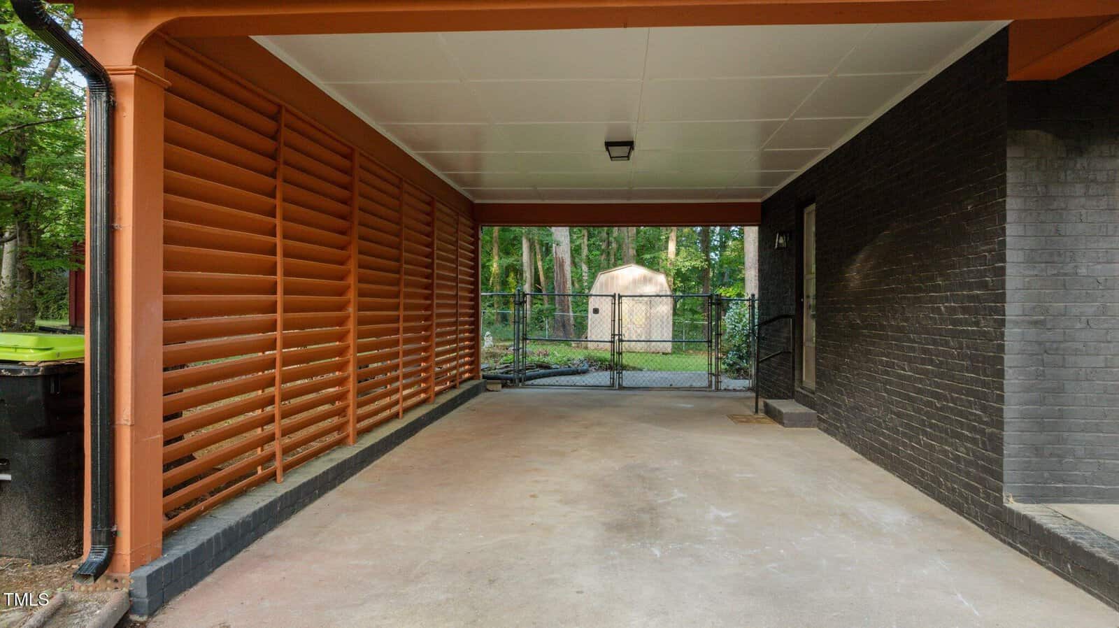 A covered carport with a concrete floor, orange support beams, slatted wood privacy screen on one side, black brick wall on the other, and a chain-link gate at the back opening to a yard with trees and a small shed.