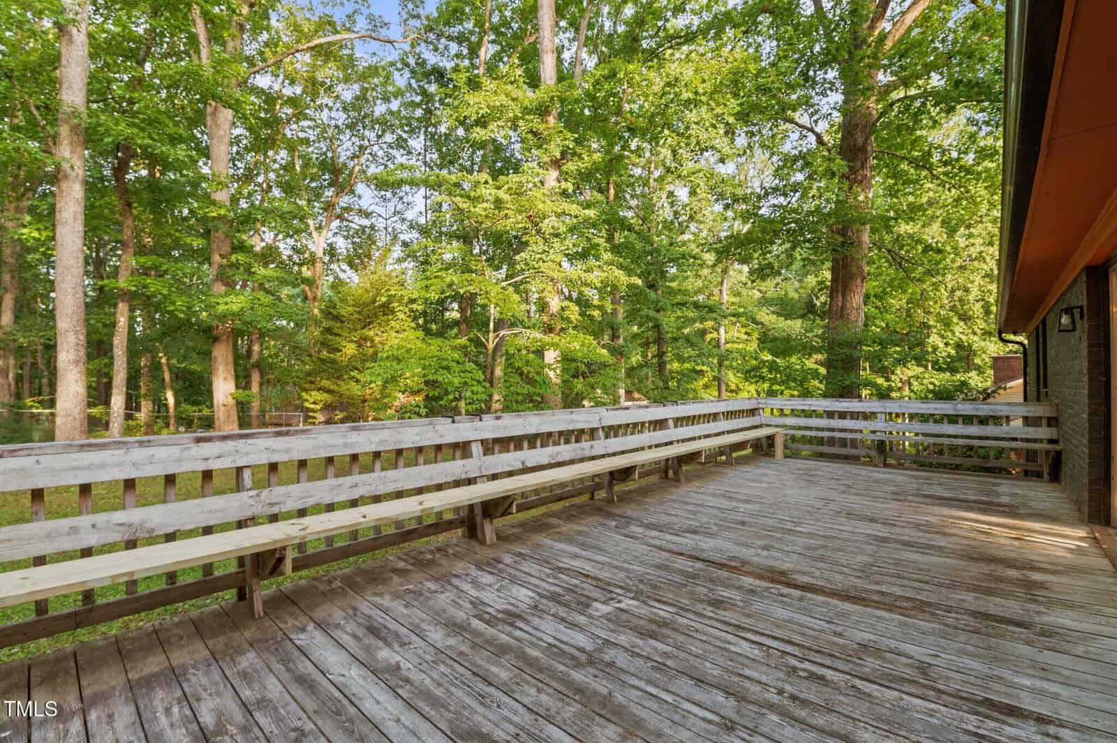 A large wooden deck with built-in benches is attached to a house, overlooking a wooded area with tall green trees. The weathered deck boards suggest outdoor use and exposure to the elements.