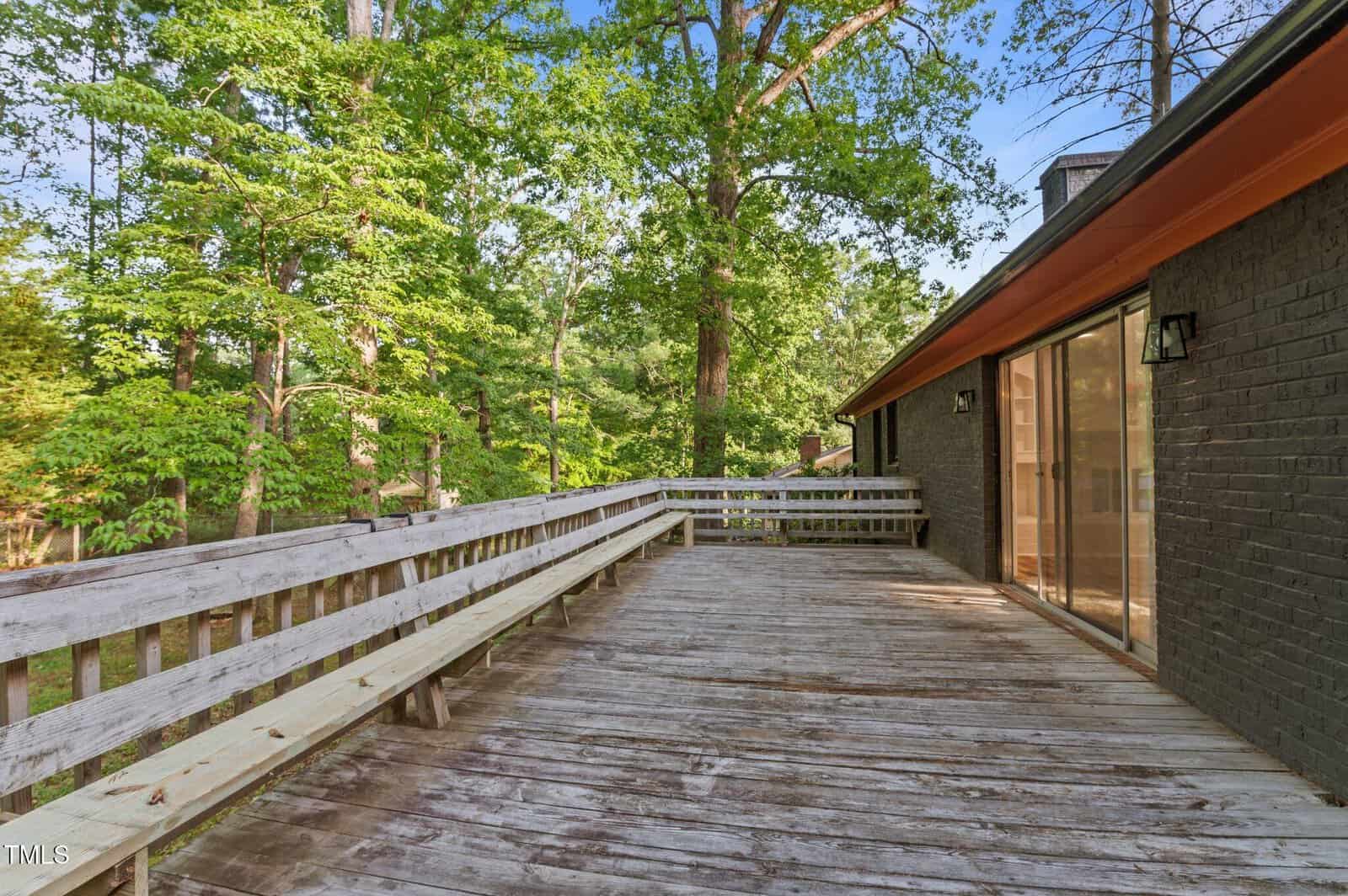 A spacious wooden deck with built-in benches runs alongside a dark brick house with large sliding glass doors. Mature trees with green leaves surround the area, creating a wooded backdrop.
