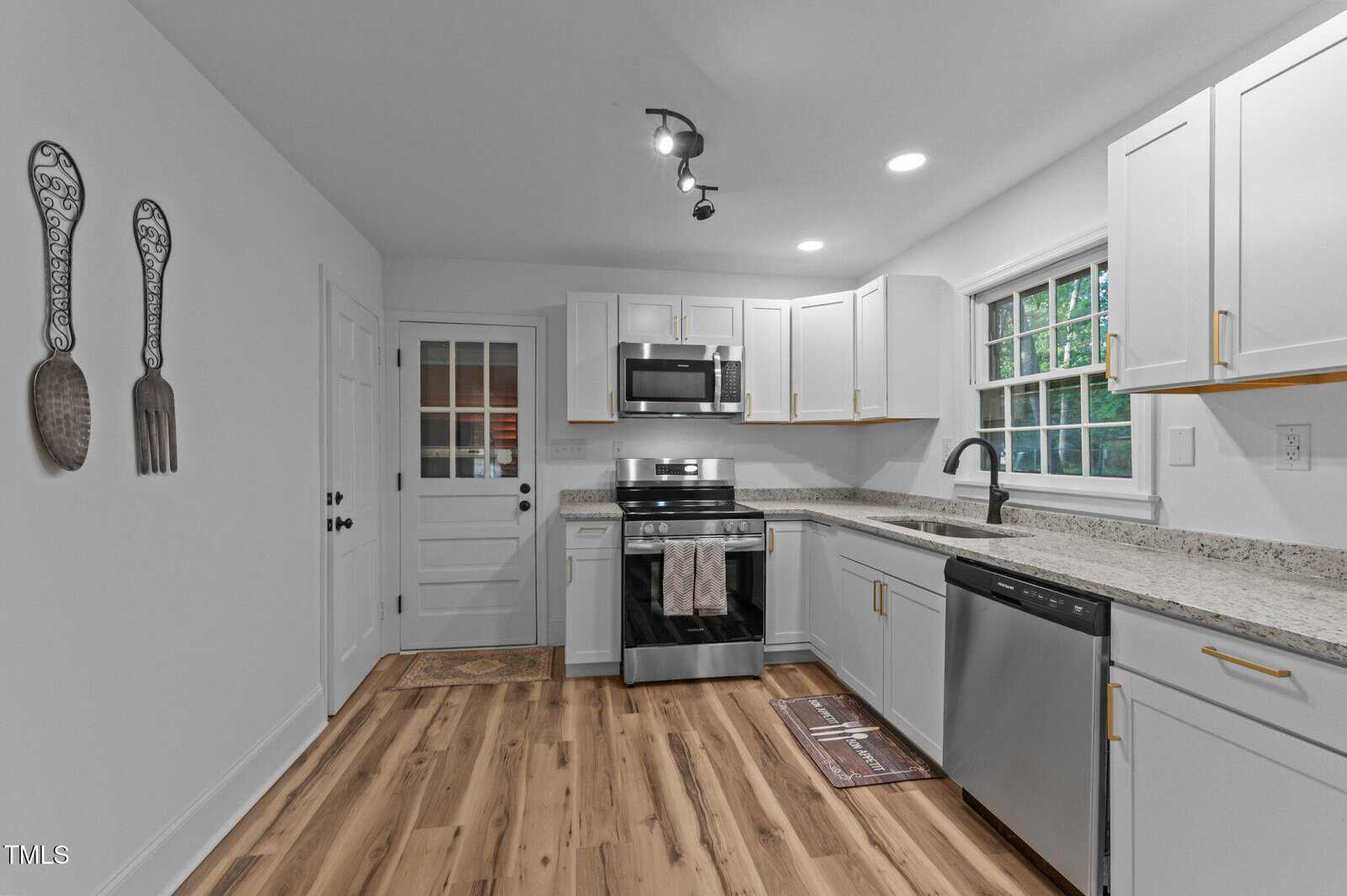 A modern kitchen with white cabinets, stainless steel appliances, light wood flooring, and granite countertops. There is a window above the sink and wall art shaped like a fork and spoon on the left wall.