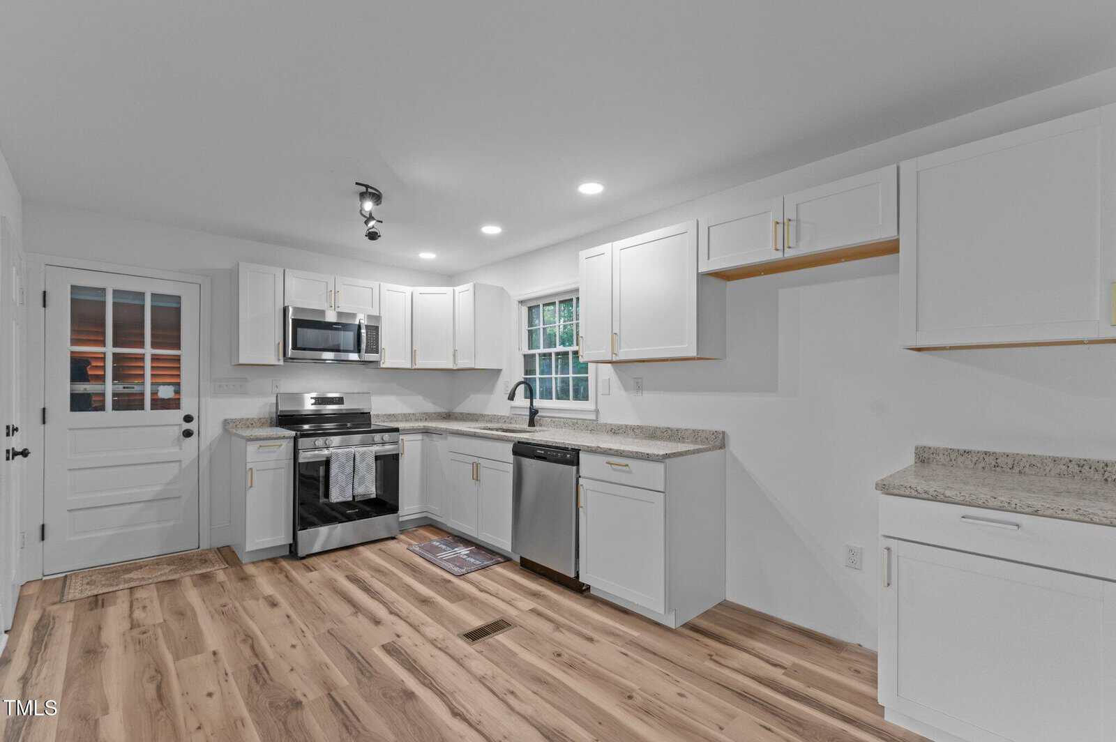 A modern kitchen with white cabinets, stainless steel appliances, granite countertops, wood flooring, a window above the sink, and a door leading outside. Ceiling lights and natural light brighten the room.