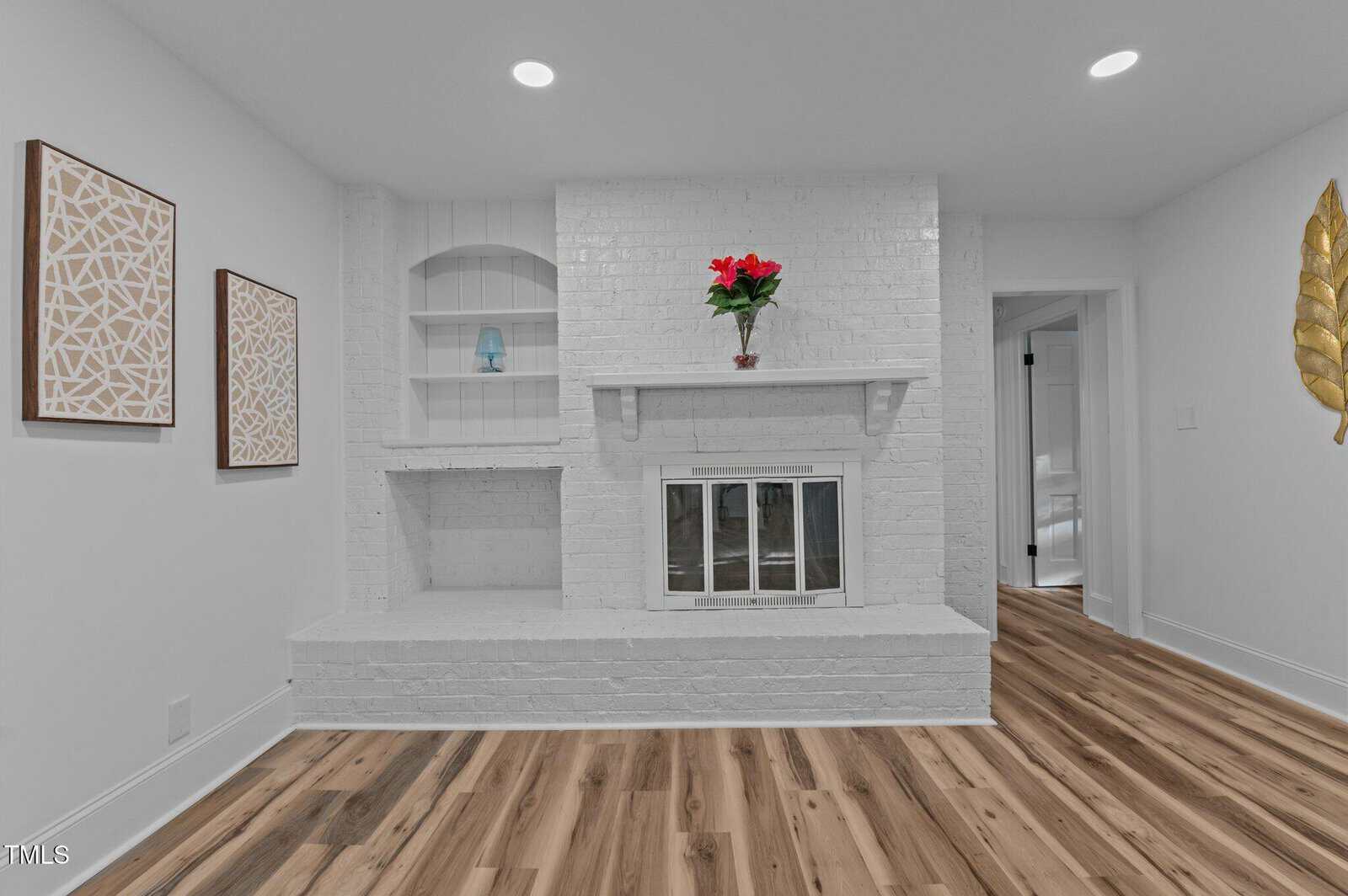 A white brick fireplace with a mantel holding a red potted plant is set against a white wall. Built-in shelves and two framed geometric artworks are on the left. The floor is light wood, and a gold leaf decor is on the right wall.