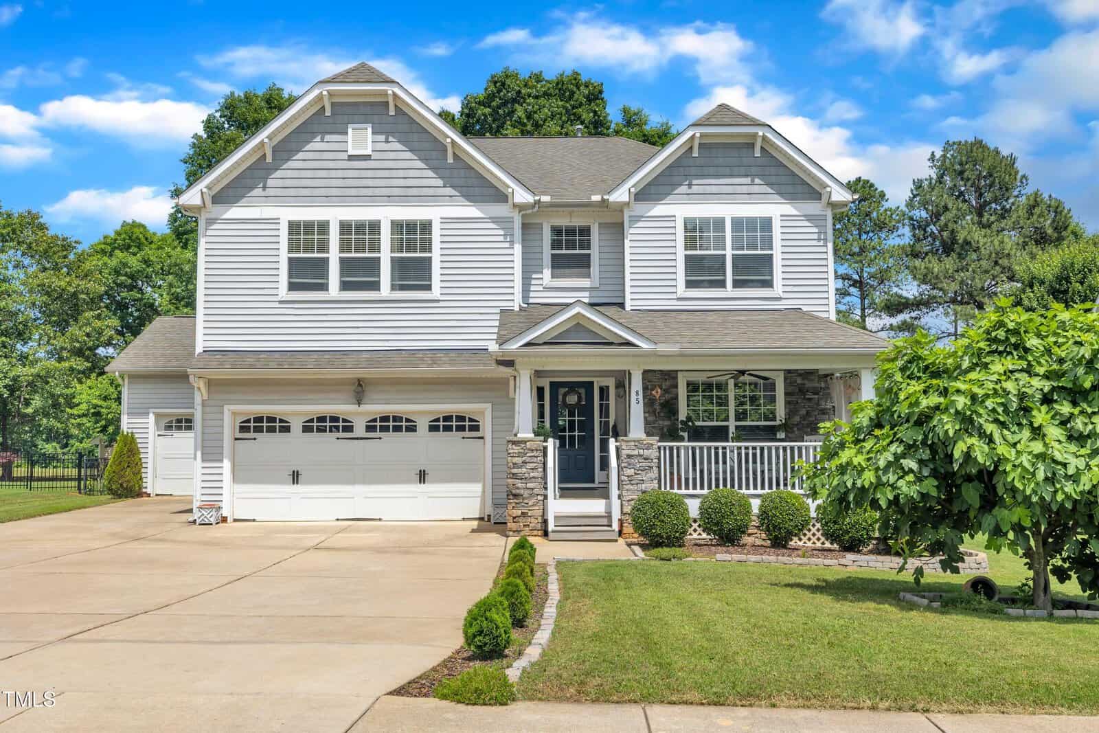 Two-story grey and white house with a double garage, front porch, and manicured lawn. The driveway is wide, and there are shrubs and trees surrounding the home under a blue sky with white clouds.