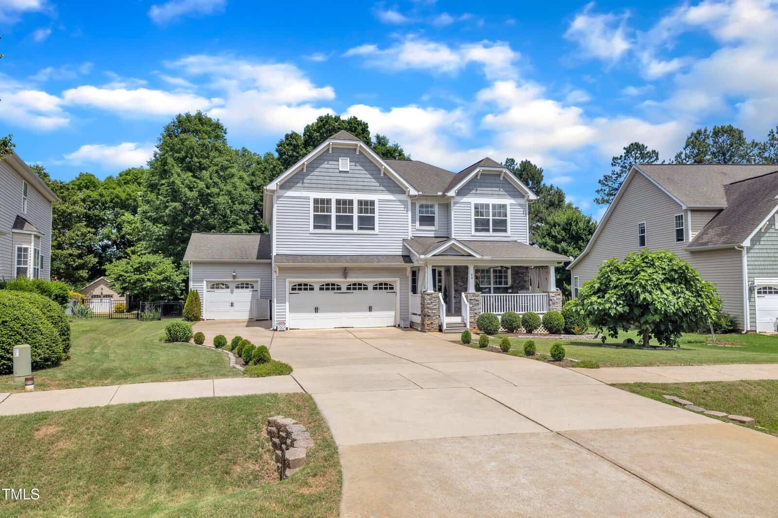 A two-story gray suburban house with white trim, a covered porch, three-car garage, and well-kept lawn, set on a sunny day with partly cloudy blue skies and neighboring homes visible on both sides.