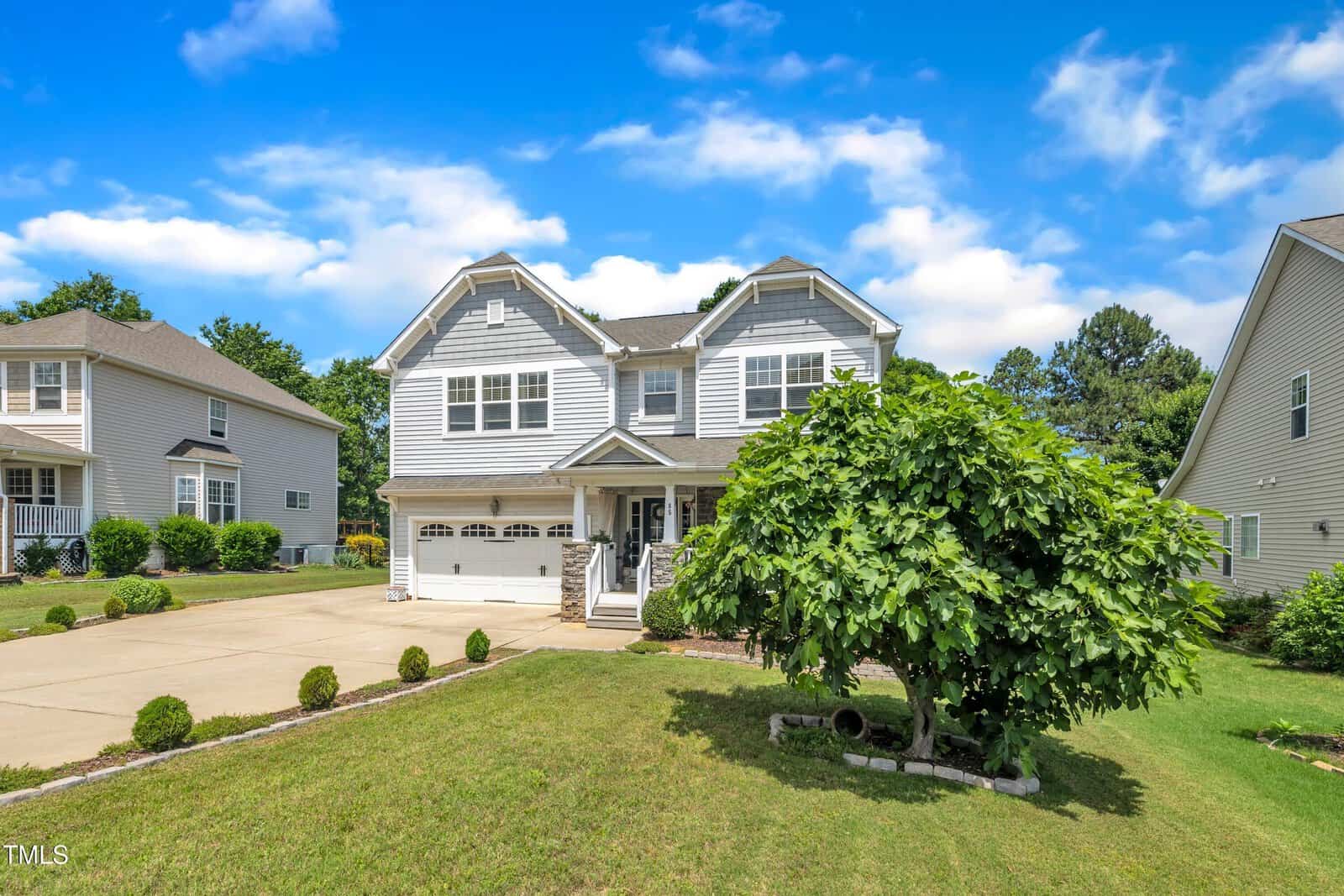 Two-story gray suburban house with white trim, double garage, and a covered porch. The front yard has a manicured lawn, a trimmed tree, and neatly edged shrubs. The sky above is bright blue with scattered clouds.