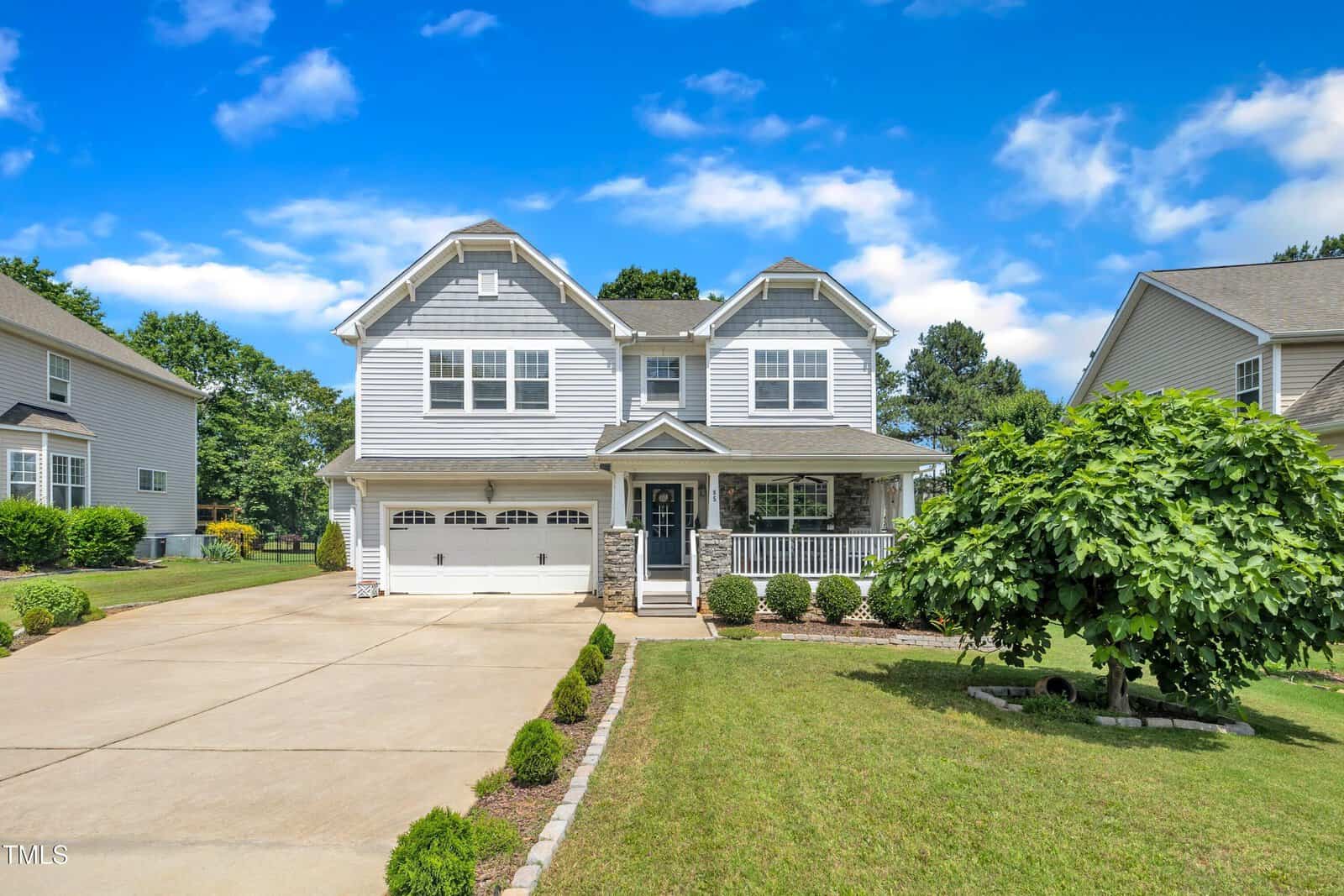 Two-story suburban house with gray siding, white trim, and a covered front porch. There is a double garage, a manicured lawn, shrubs, and a small tree in the front yard under a blue sky with scattered clouds.