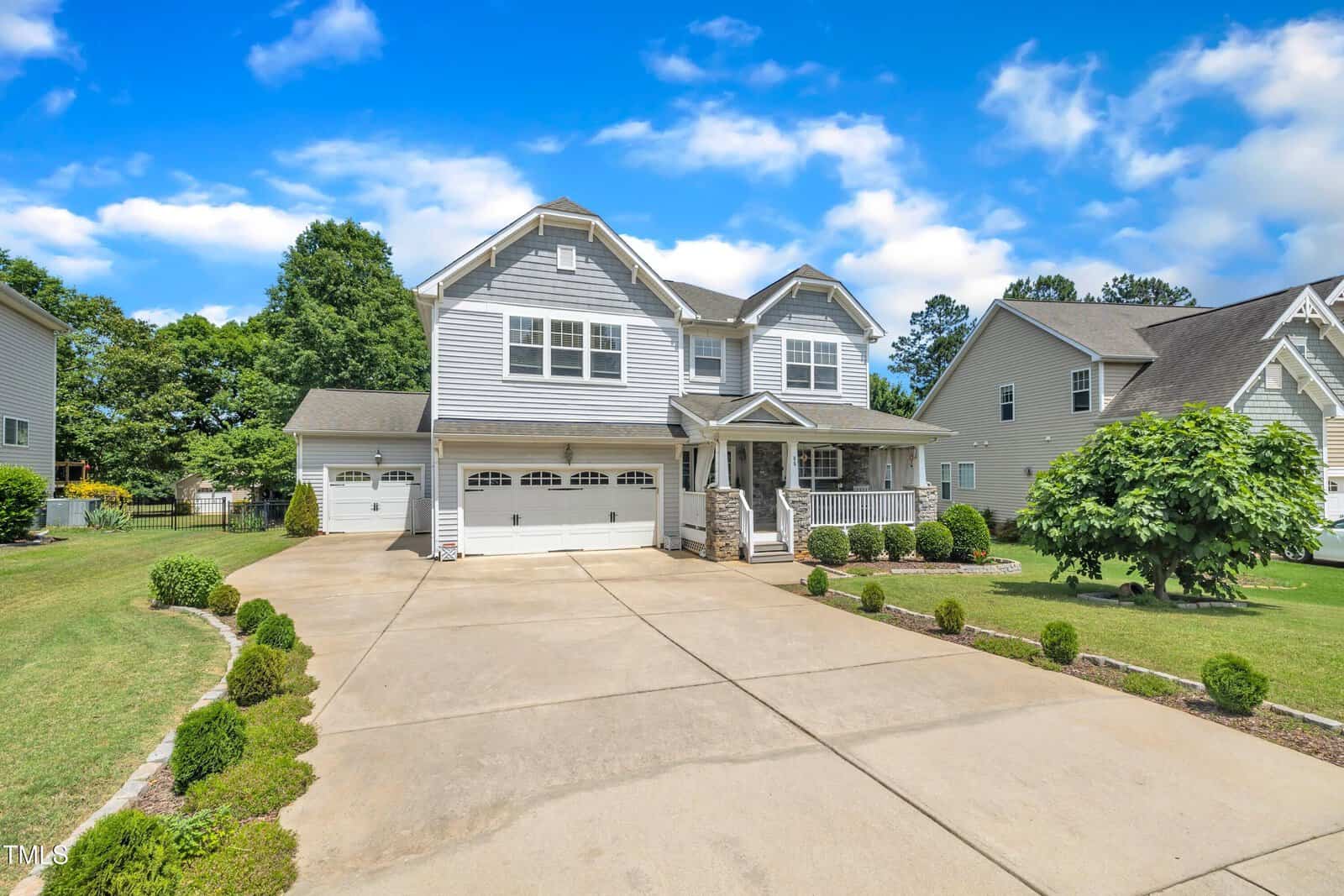 A large two-story suburban house with gray siding, white trim, and a three-car garage. The driveway is wide and bordered by manicured shrubs, with a well-kept lawn and neighboring houses visible.