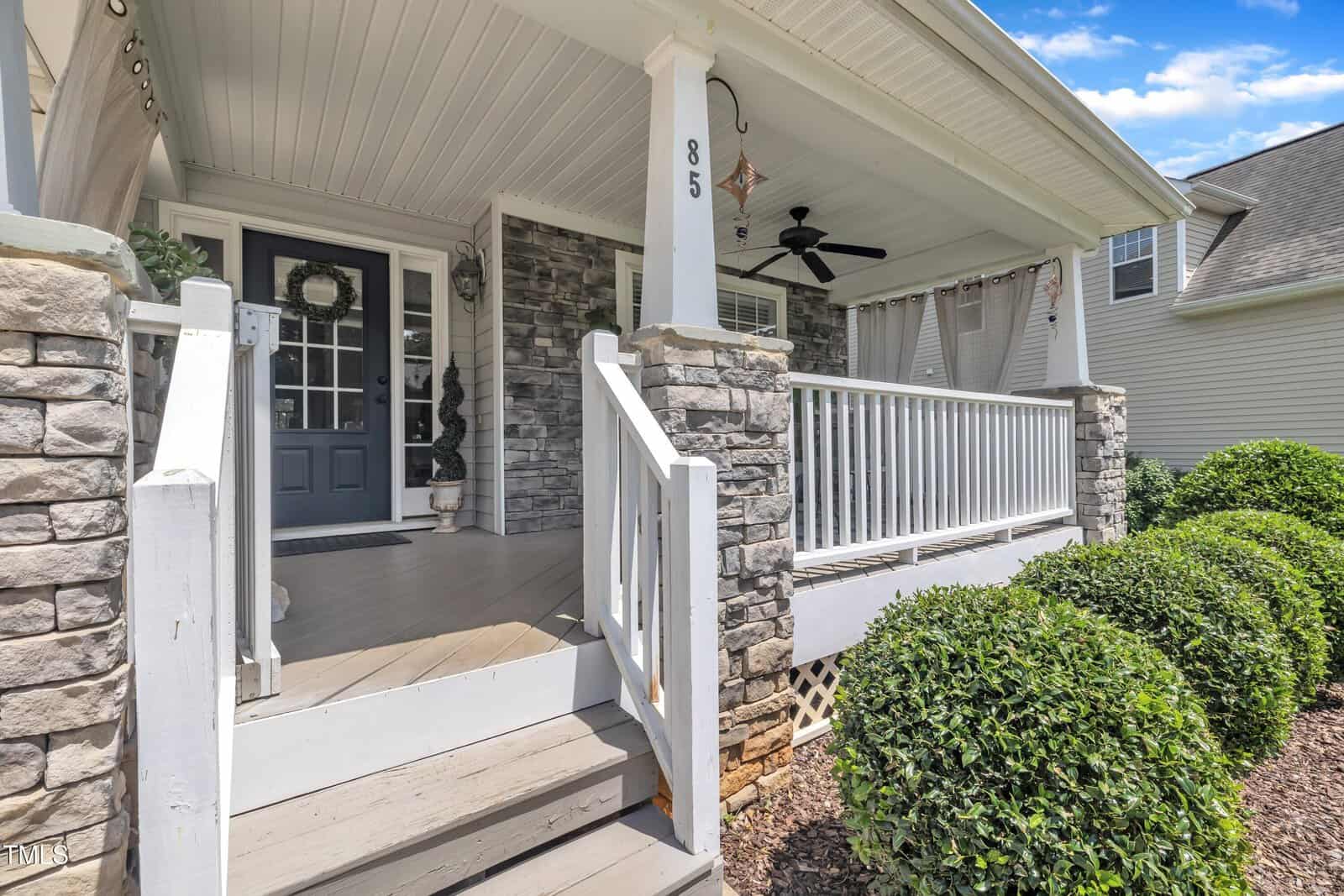 Front view of a house porch with stone columns, white railing, ceiling fan, hanging curtains, and a gray door. Four steps lead up to the porch, surrounded by trimmed bushes and mulch. House number 85 is visible on a column.