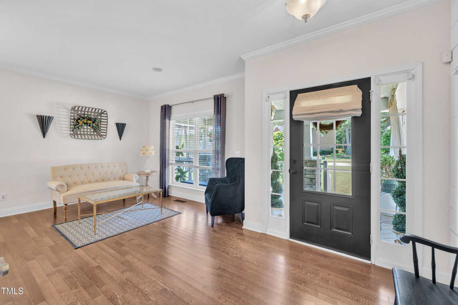 Bright living room with hardwood floors, cream walls, a glass-paned black front door, beige tufted sofa, gold coffee table, patterned rug, black armchair, and large windows with sheer and dark curtains.