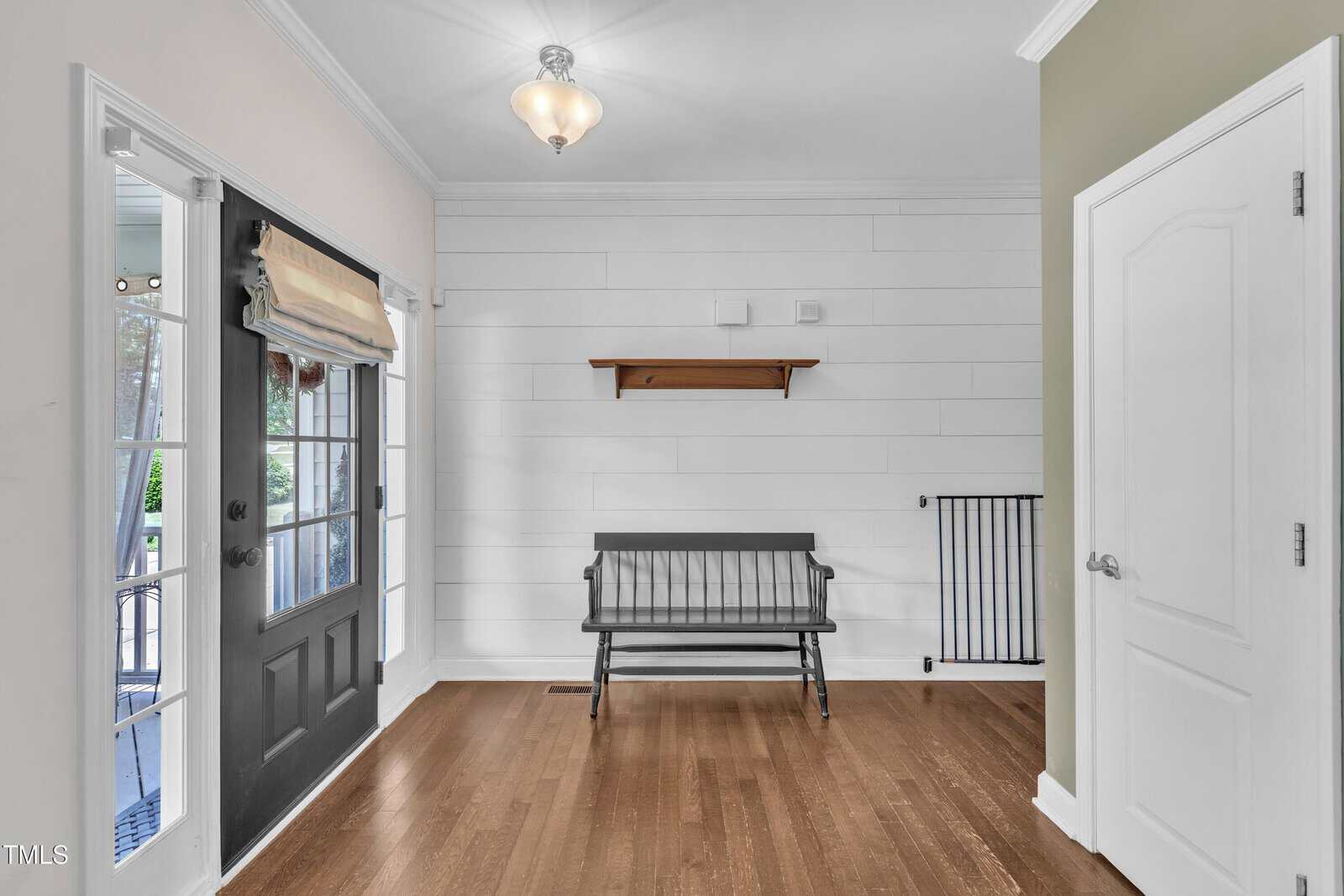Entryway with a black door, a window with a beige Roman shade, a black wooden bench, a floating wooden shelf above, white shiplap wall, hardwood floor, and a closed white door on the right.
