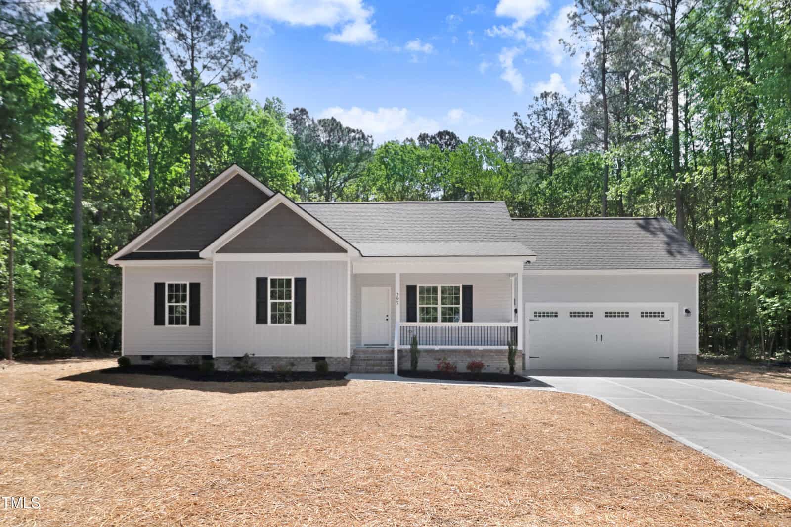 Single-story white house with dark gray accents, a front porch, attached two-car garage, and concrete driveway, set against a backdrop of tall green trees and clear blue sky.