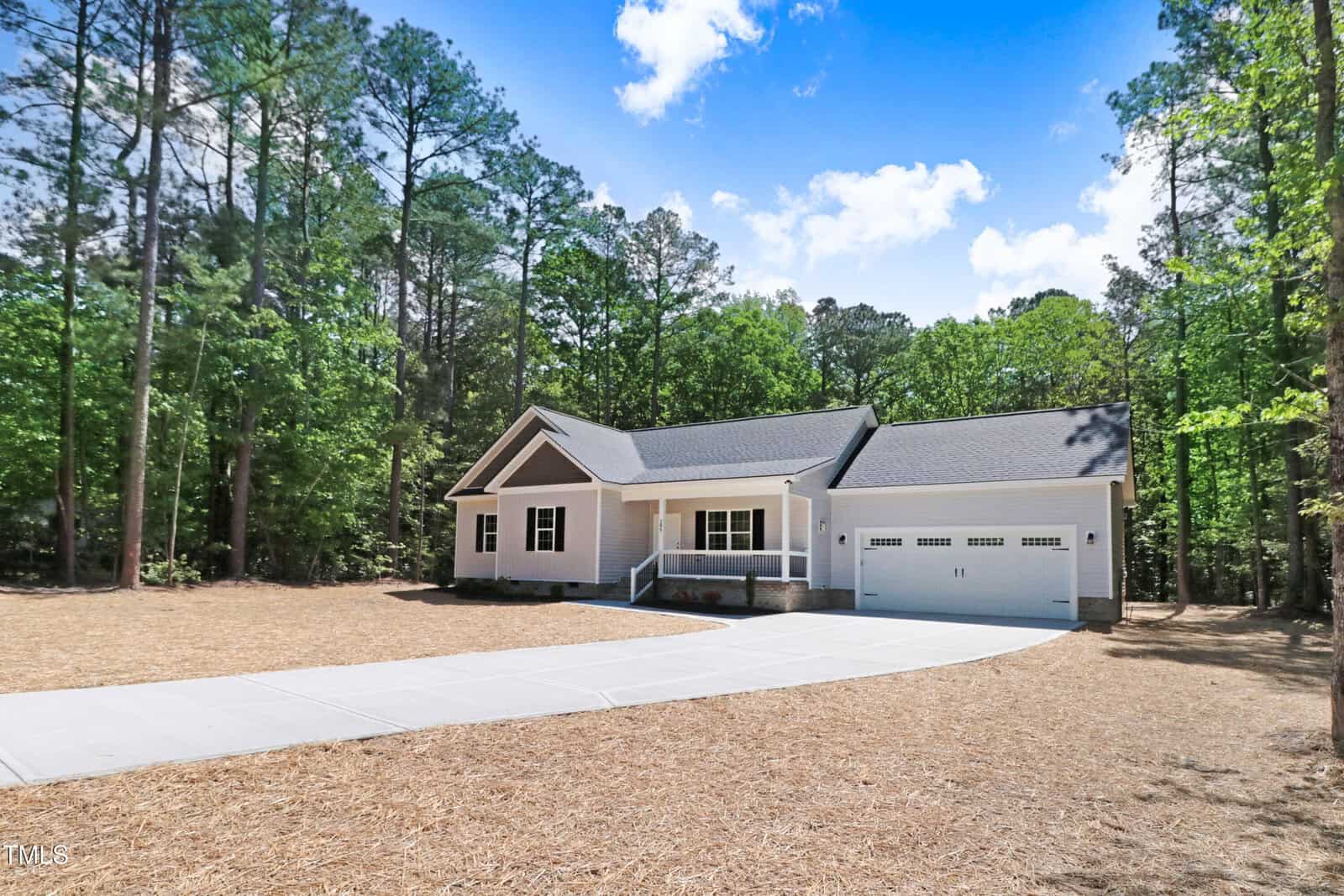 A single-story house with a front porch, two-car garage, and gray siding sits among tall pine trees. A wide concrete driveway leads up to the house, and the yard is covered with straw. The sky is partly cloudy.