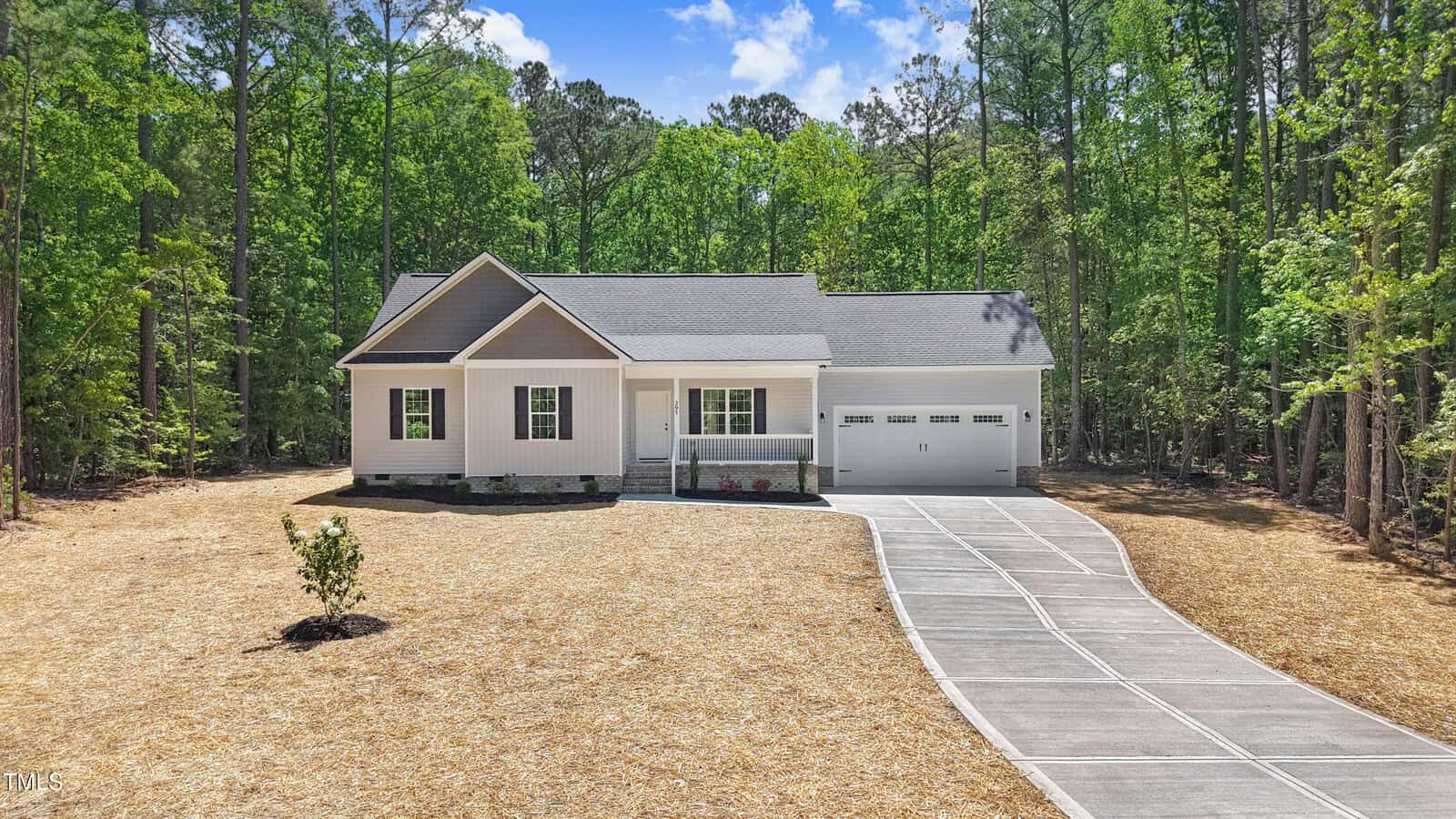 A white single-story house with a front porch, attached two-car garage, and a long concrete driveway, surrounded by tall trees and set against a forested background under a blue sky.