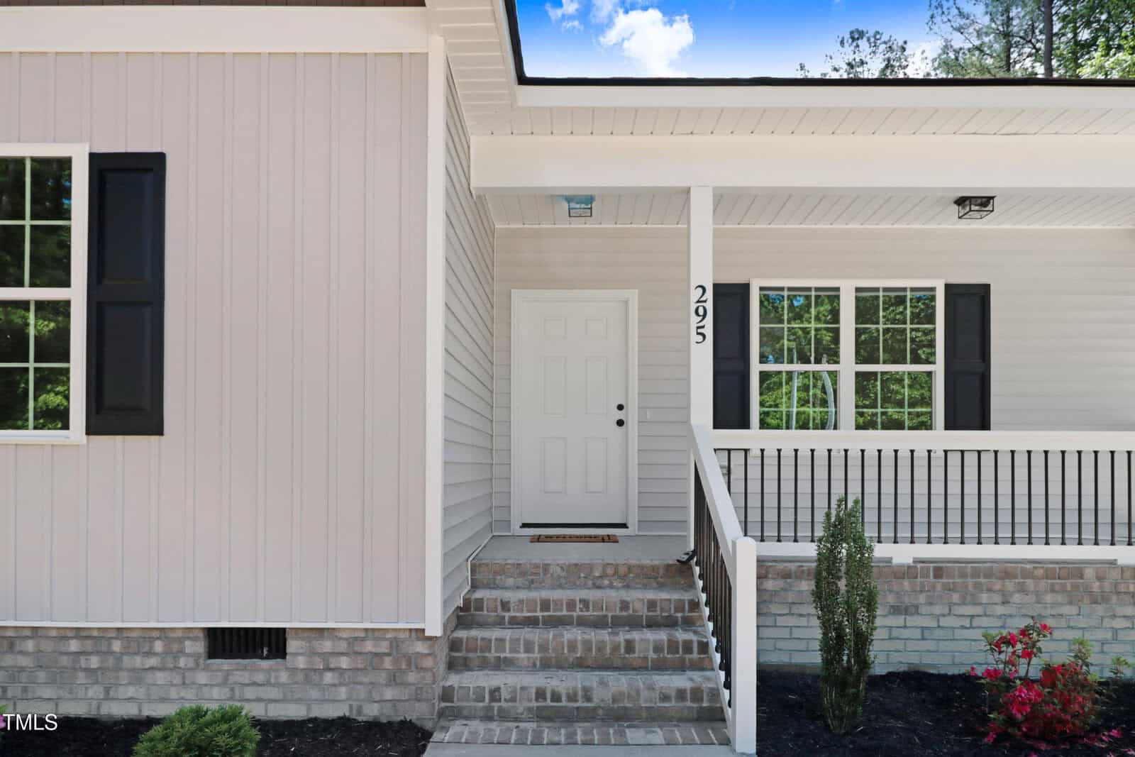 Front entrance of a house with beige siding, white door, black window shutters, a covered porch with railings, brick steps, and small plants along the walkway. The house number "295" is displayed beside the door.