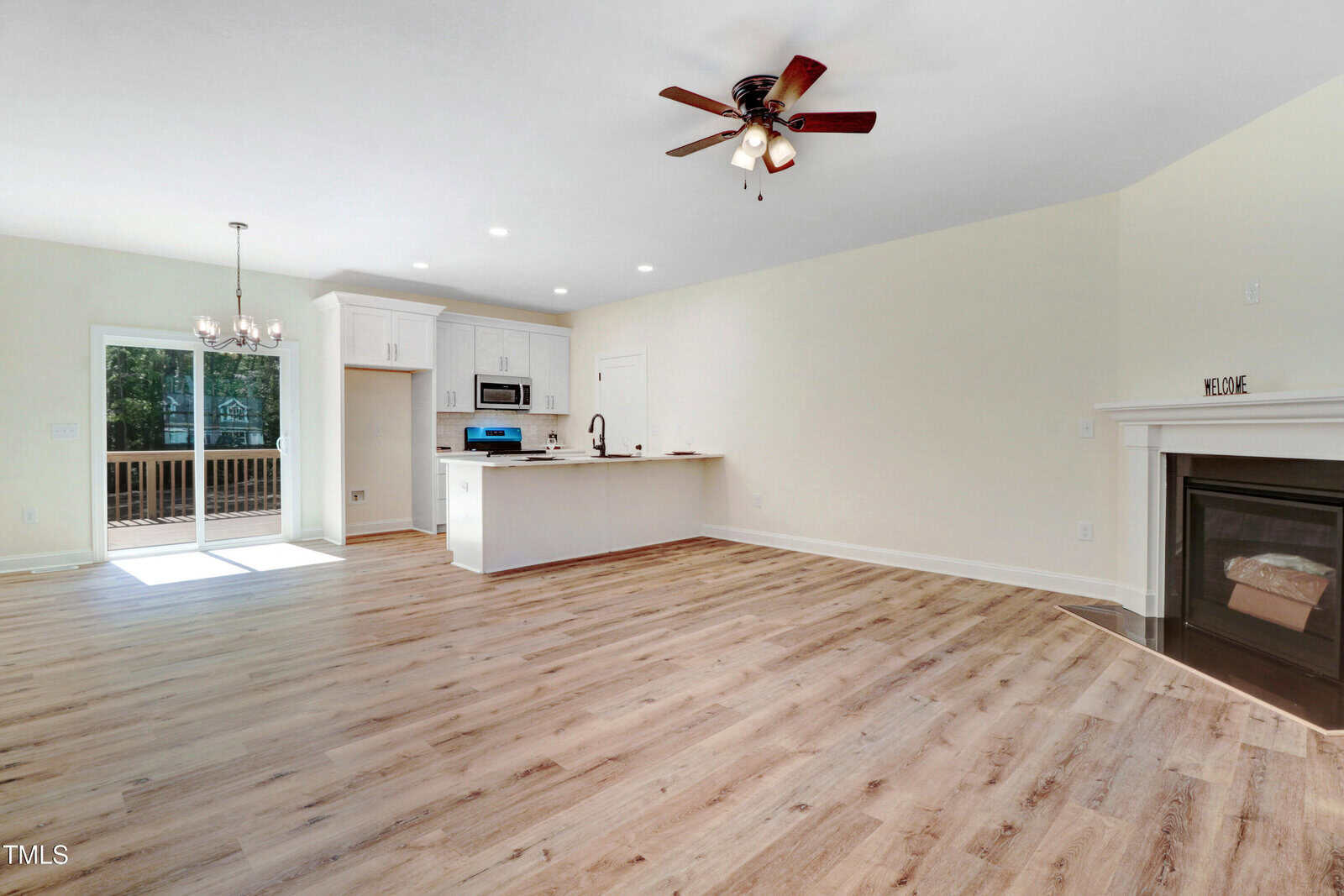 Open-concept living area with wood-style flooring, a kitchen with white cabinets and stainless steel appliances, an island with a sink, a fireplace, ceiling fan, chandelier, and sliding doors to a deck.