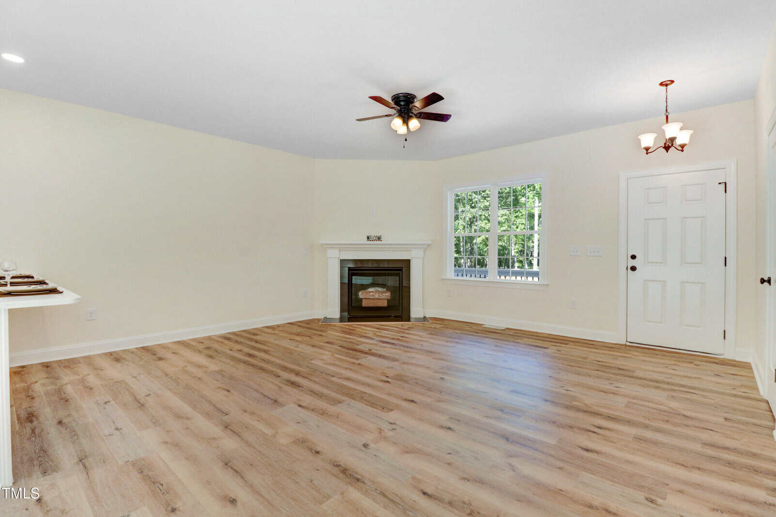 Unfurnished living room with light wood flooring, cream walls, a ceiling fan, fireplace, large window, and a door next to a small chandelier. Natural light fills the space.