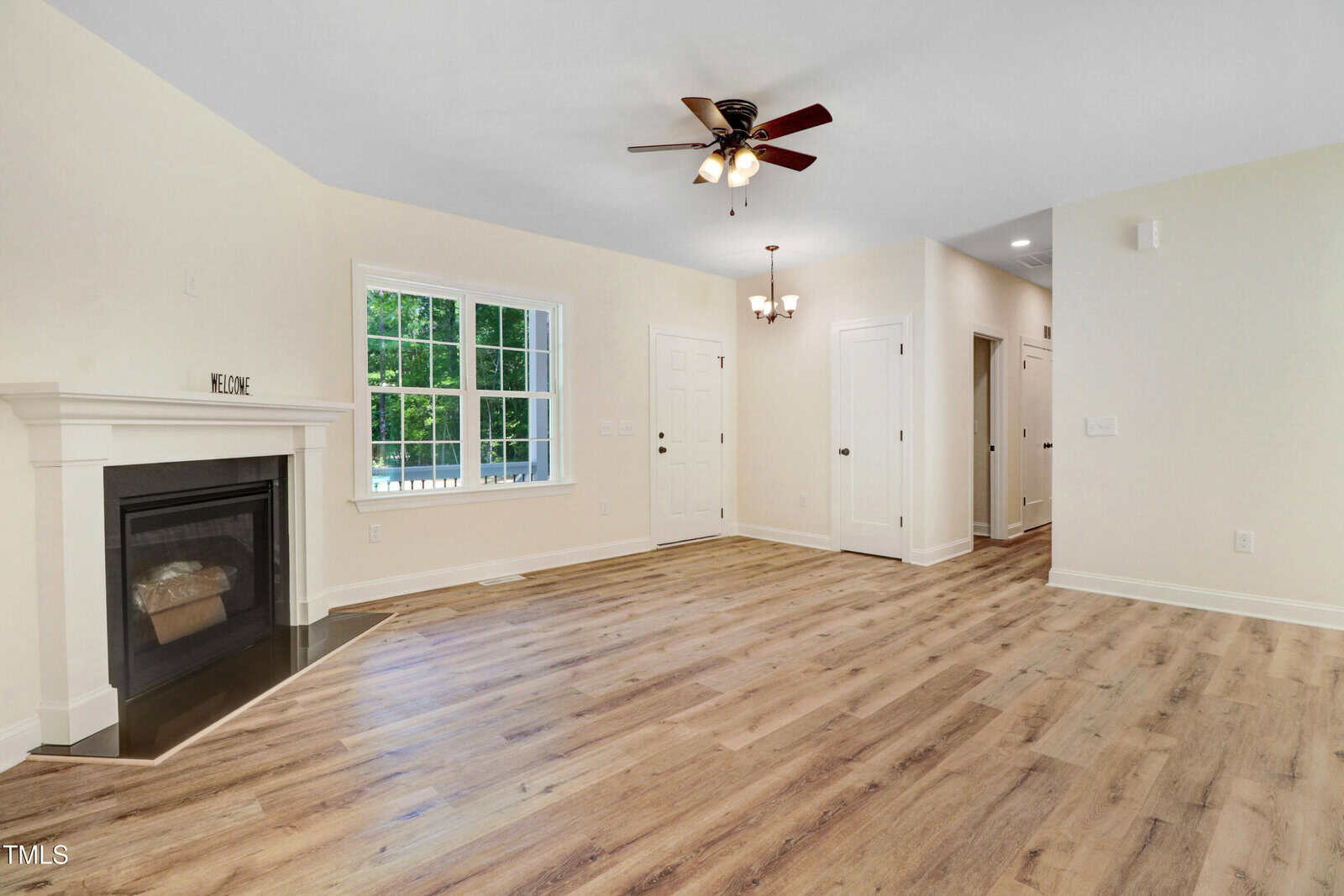 A spacious, empty living room with light wood flooring, a ceiling fan, a fireplace on the left, large window, and multiple white doors leading to other rooms. Walls are cream-colored and lighting fixtures are visible.