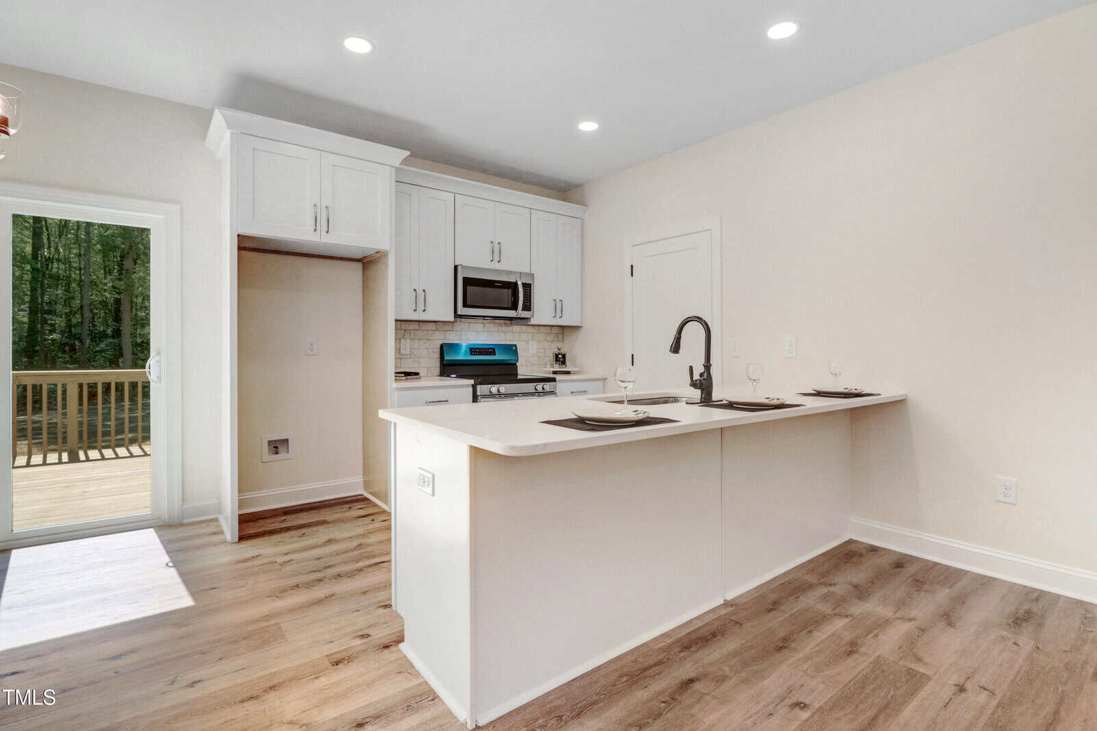 Modern kitchen with white cabinets, stainless steel appliances, a center island with a sink and faucet, two place settings, wood flooring, and a sliding glass door leading to a wooden deck outside.