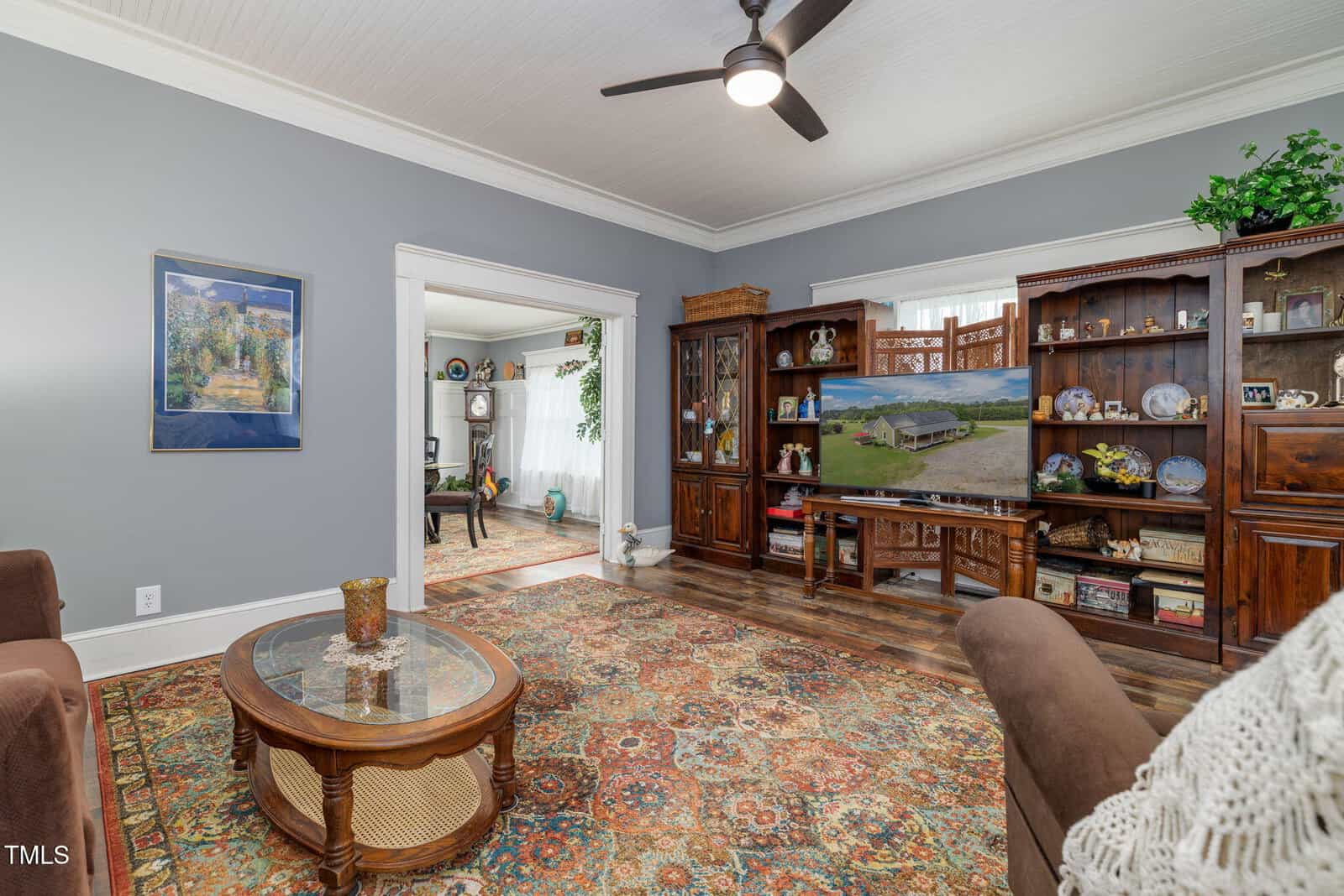 A living room with a patterned rug, brown sofa, glass coffee table, wooden shelves displaying decor, a ceiling fan, and a large doorway leading to another room with light curtains and additional furniture.
