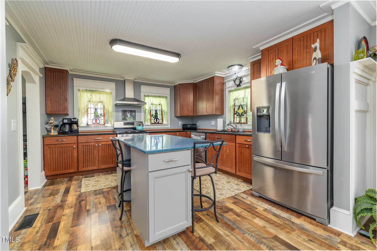 A modern kitchen with wooden cabinets, stainless steel appliances, a center island with two chairs, a gray countertop, and patterned hardwood flooring. Two windows with green curtains are above the sink area.