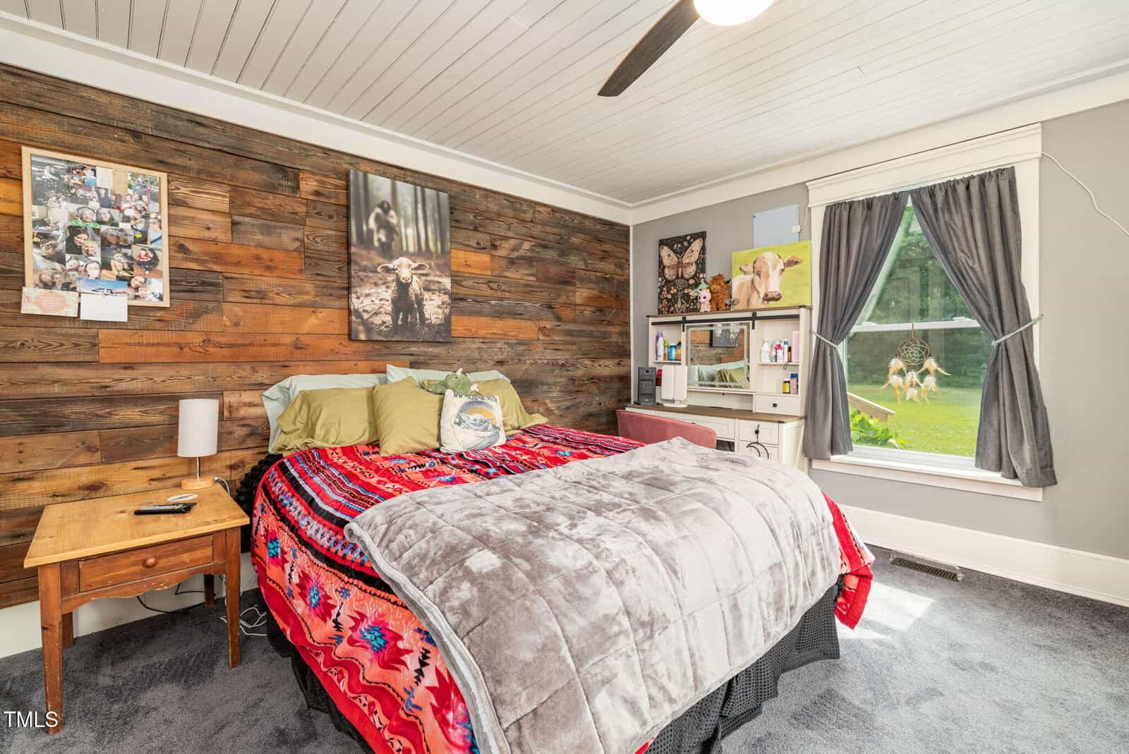 Cozy bedroom with a rustic wooden accent wall, a bed with colorful blankets and pillows, a wooden nightstand, and a window with gray curtains showing a green yard outside. Art and animal photos decorate the walls.