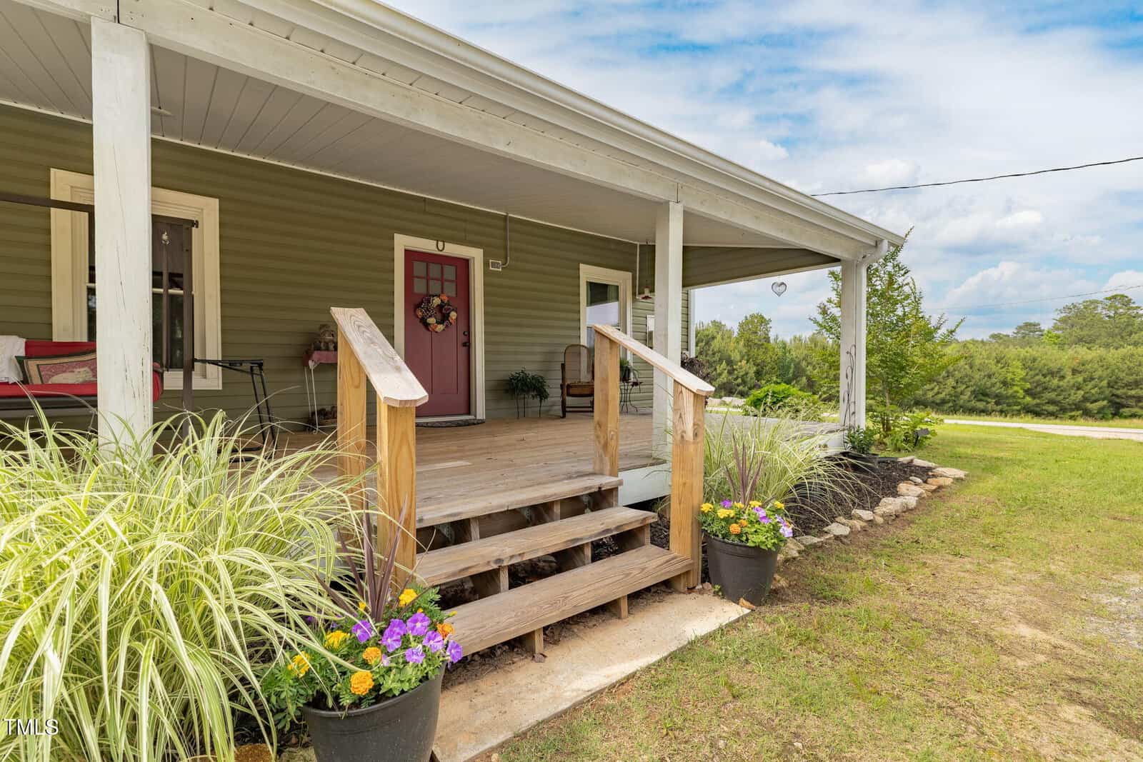 A cozy front porch with wooden steps, potted flowers, a red front door with a wreath, and a bench. The house has green siding and white trim, and the yard is grassy with shrubs and trees in the background.