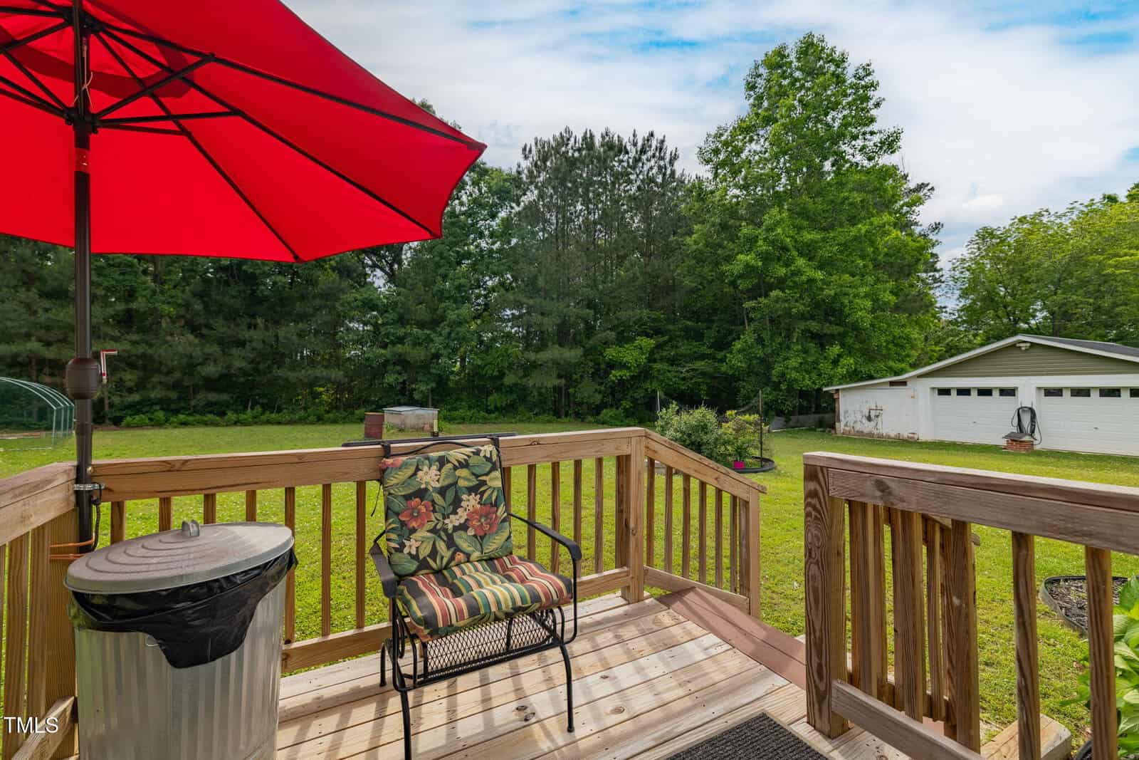 A wooden deck with a floral cushioned chair, red umbrella, and trash can overlooks a grassy yard with trees and a white detached garage in the background under a partly cloudy sky.