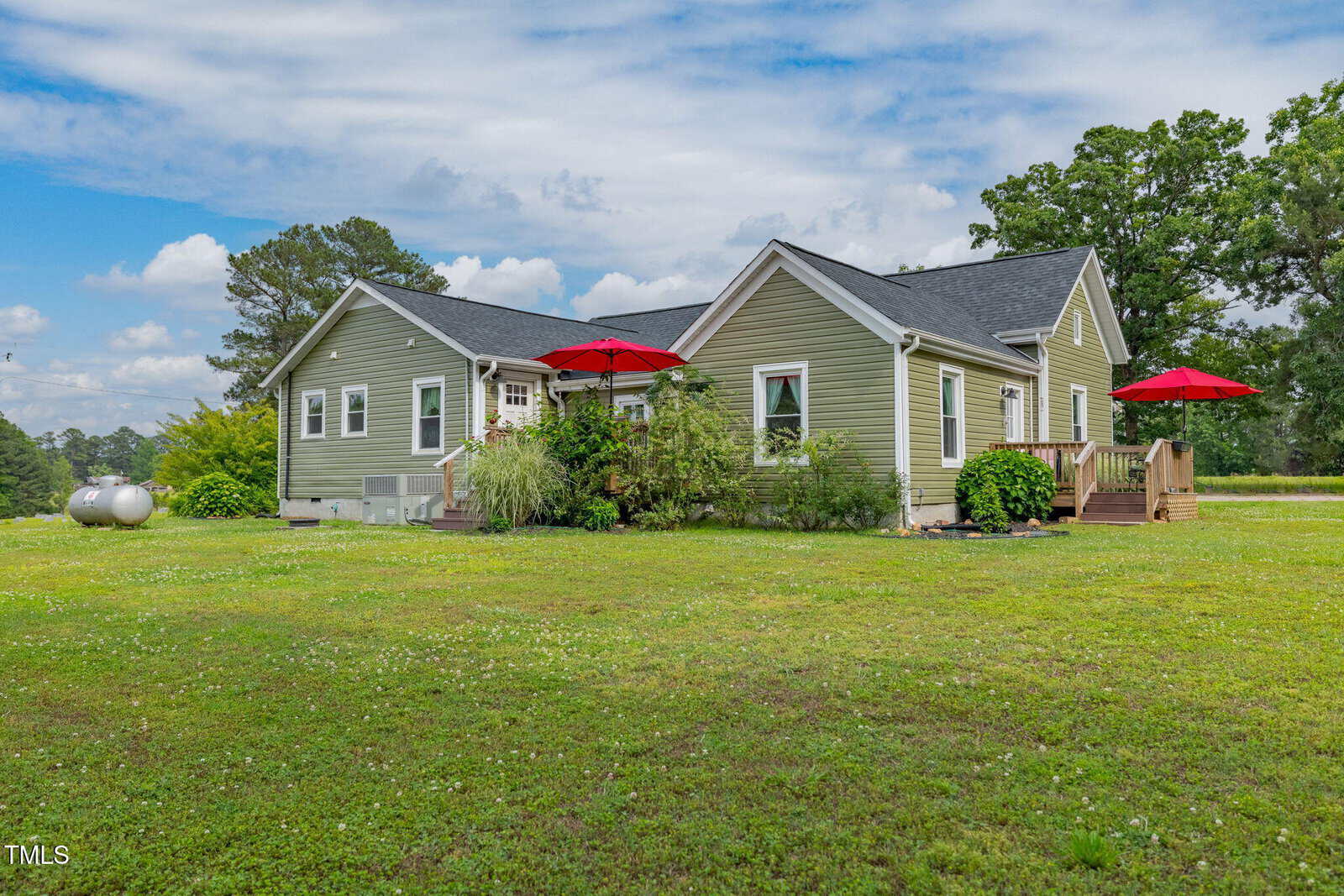 Single-story green house with white trim, surrounded by a large grassy yard. There are two red patio umbrellas, one on the deck and one near the side door, and propane tanks visible on the left side of the house.