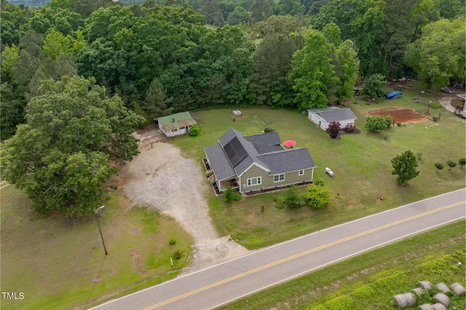 Aerial view of a single-story house with a gray roof, gravel driveway, small outbuildings, and scattered trees, located next to a two-lane road with green forest in the background.