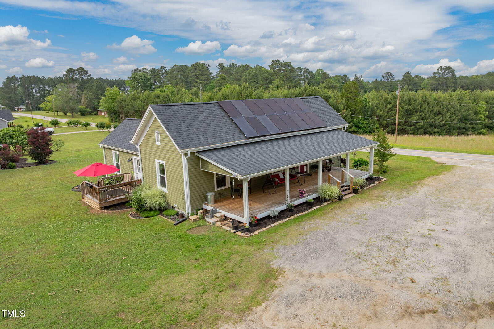 A green house with a front porch and solar panels on the roof sits on a spacious grassy lot, with a deck and red umbrella on one side and surrounded by trees under a partly cloudy sky.