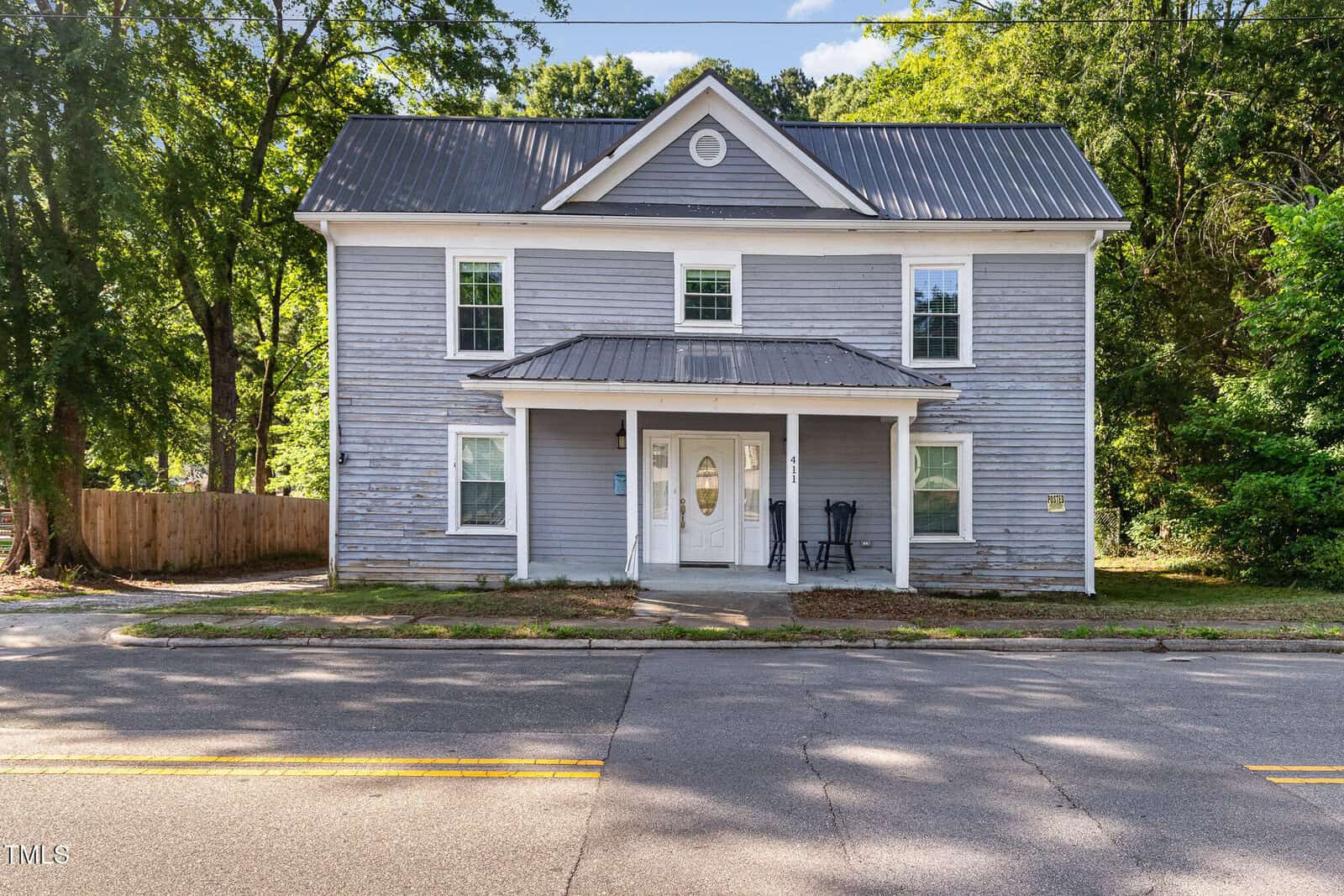 A two-story gray house with white trim and a metal roof, featuring a small covered porch with double doors and two chairs, sits along a quiet street surrounded by trees and a wooden fence.