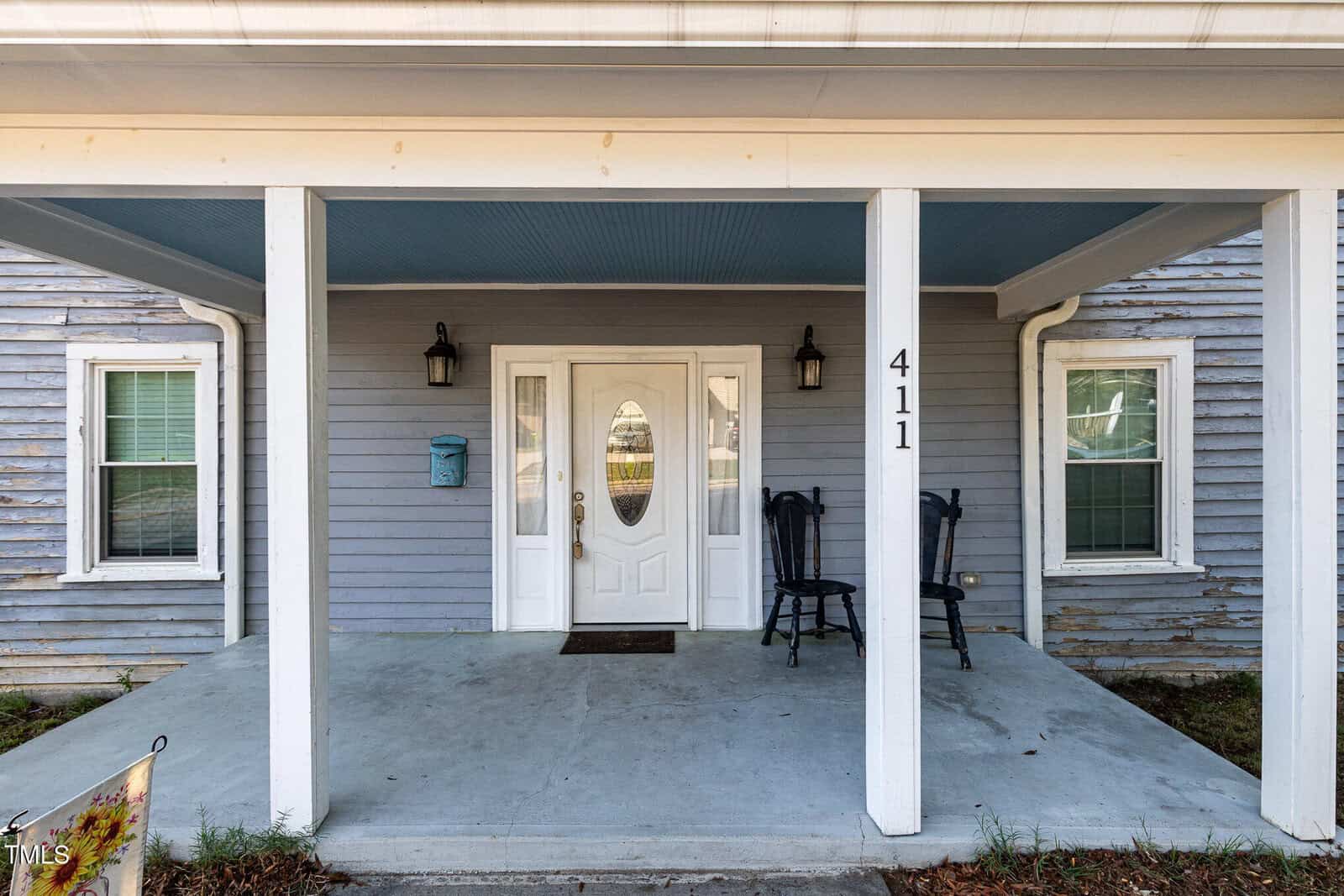 A covered front porch with two black chairs, white double front doors with an oval window, and house number 411 on a white pillar. The house has light blue siding and two windows on either side of the door.