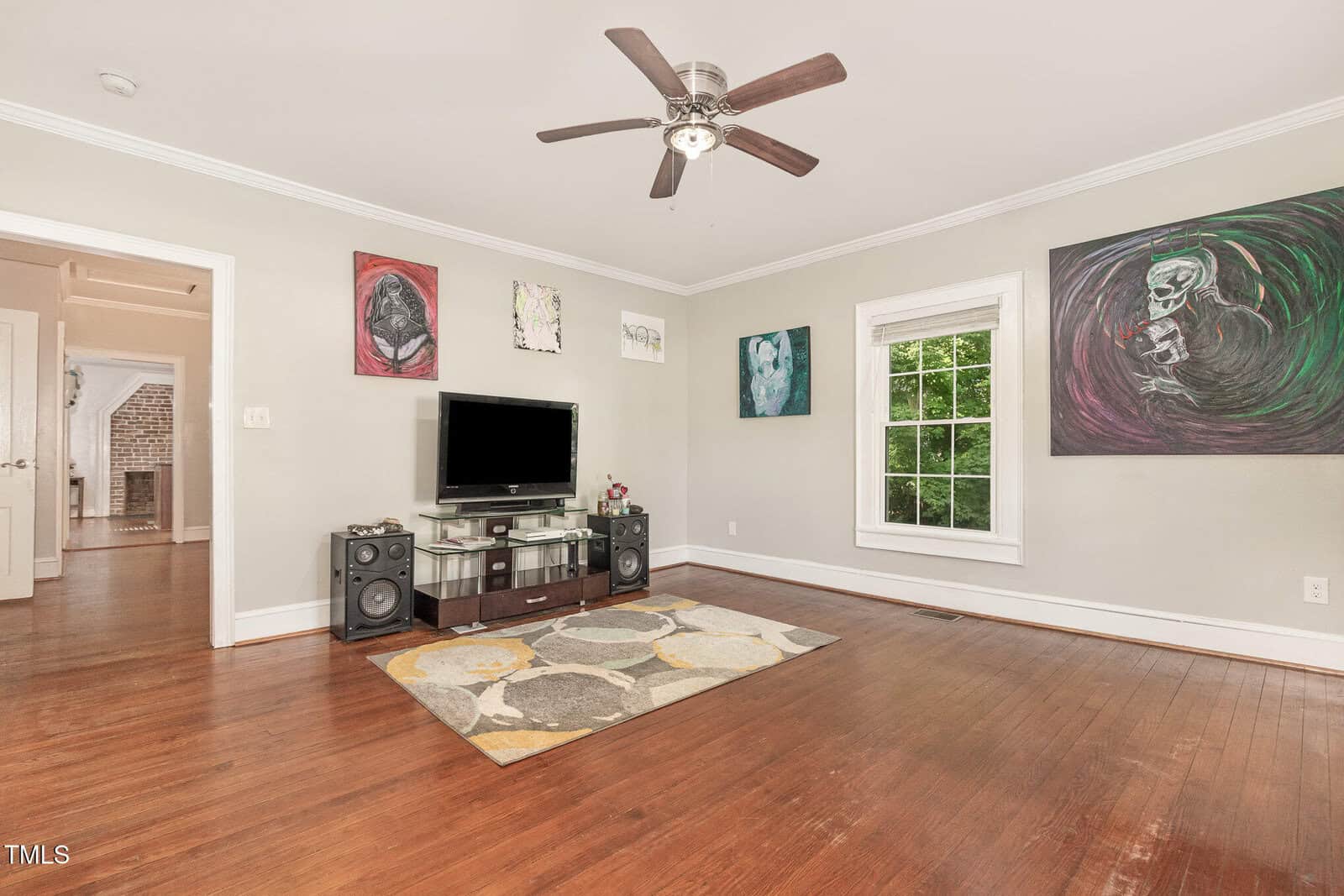 A living room with hardwood floors, a ceiling fan, a TV on a glass stand with speakers, a geometric rug, and colorful abstract art on light gray walls. A window lets in natural light and shows green trees outside.