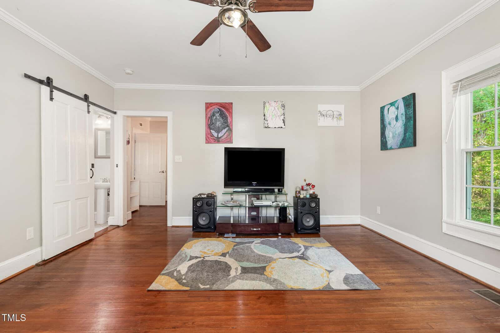 A living room with hardwood floors, a ceiling fan, a TV on a glass stand between two speakers, colorful abstract art on the walls, a patterned rug, a sliding barn door, and a large window with a view of greenery.