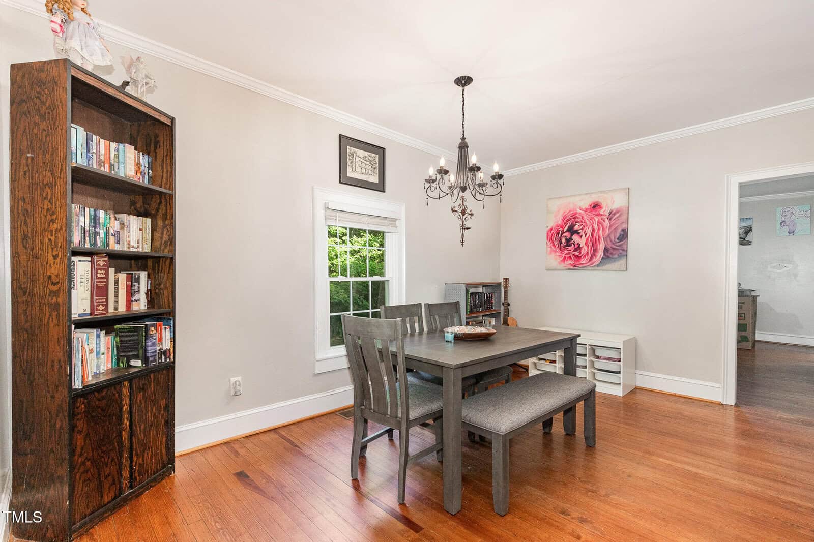 Dining room with wooden floor, bookshelf, rectangular table with four chairs and a bench, a black chandelier, a window, large pink floral artwork on the wall, and a small storage unit in the corner.