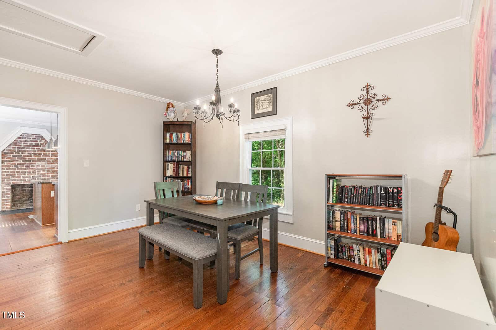 A dining room with a wooden table, four chairs, a bench, two bookshelves filled with books, a guitar, a window, chandelier, and wall decor. Hardwood floors and light-colored walls are visible.