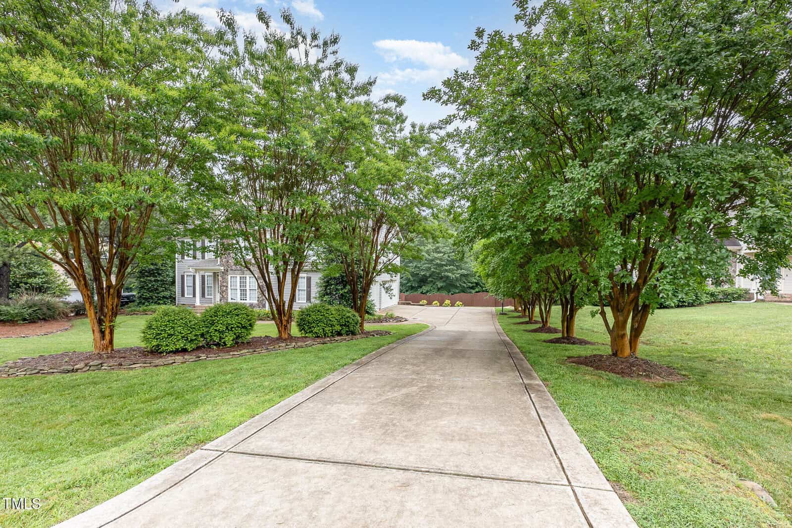 A long, curved concrete driveway leads to a house partially hidden by tall green trees and landscaped bushes on both sides, with grass lawns and a bright, partly cloudy sky.