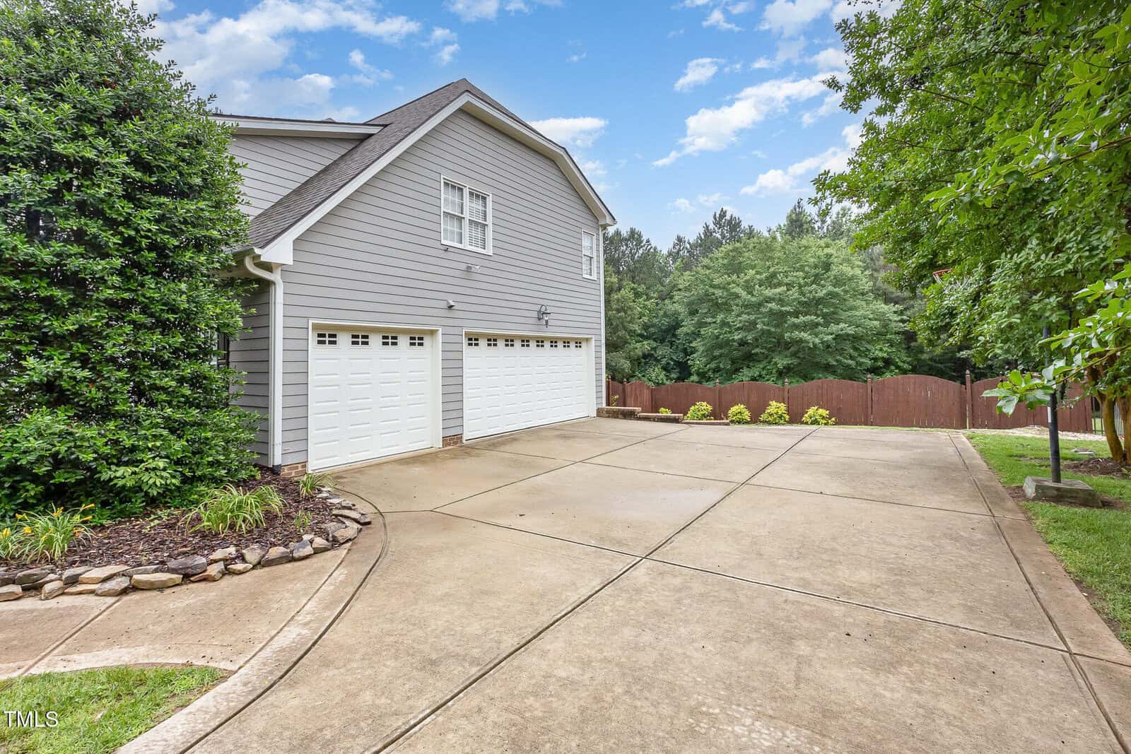 A wide concrete driveway leads to a gray house with three white garage doors, surrounded by green trees and a wooden fence, under a partly cloudy sky.