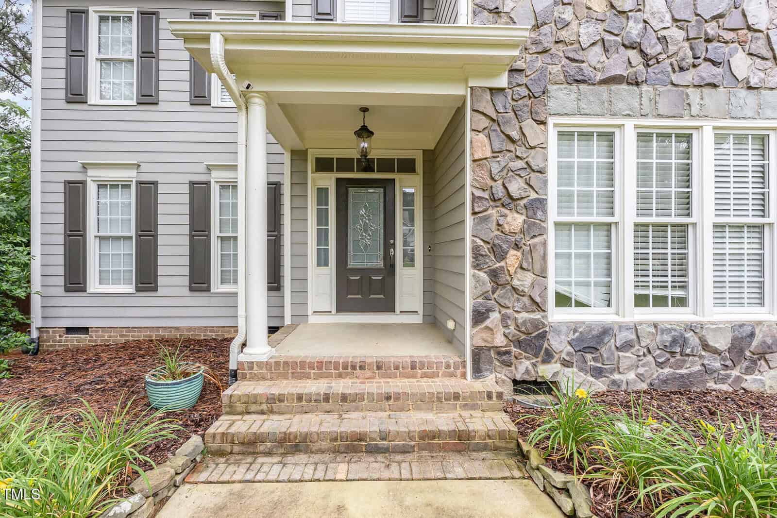 A front porch of a house with brick steps, a black front door with a decorative glass panel, white columns, gray siding with dark shutters, and a section of stone exterior. Potted plants and landscaping border the entryway.