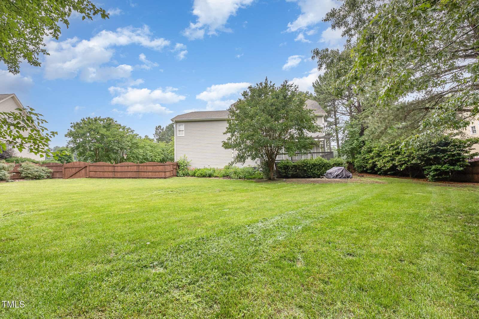 A spacious backyard with green grass, a few trees, a brown wooden fence, and a house in the background under a partly cloudy blue sky.