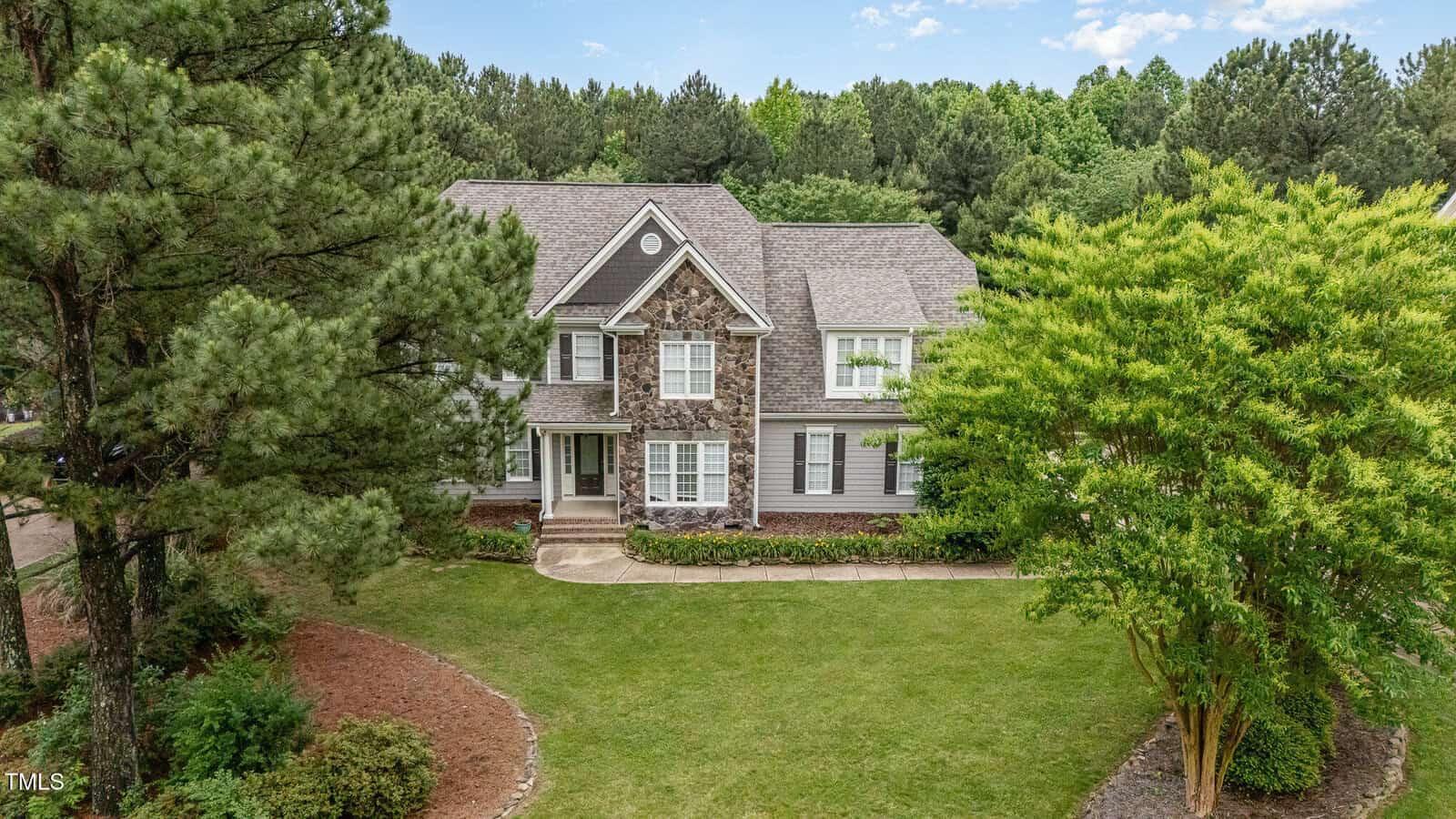 Two-story suburban house at 50 Pebble Creek Drive in Franklinton, featuring stone and gray siding exterior, surrounded by trees and landscaped lawn, set against a lush, wooded backdrop under a partly cloudy sky.