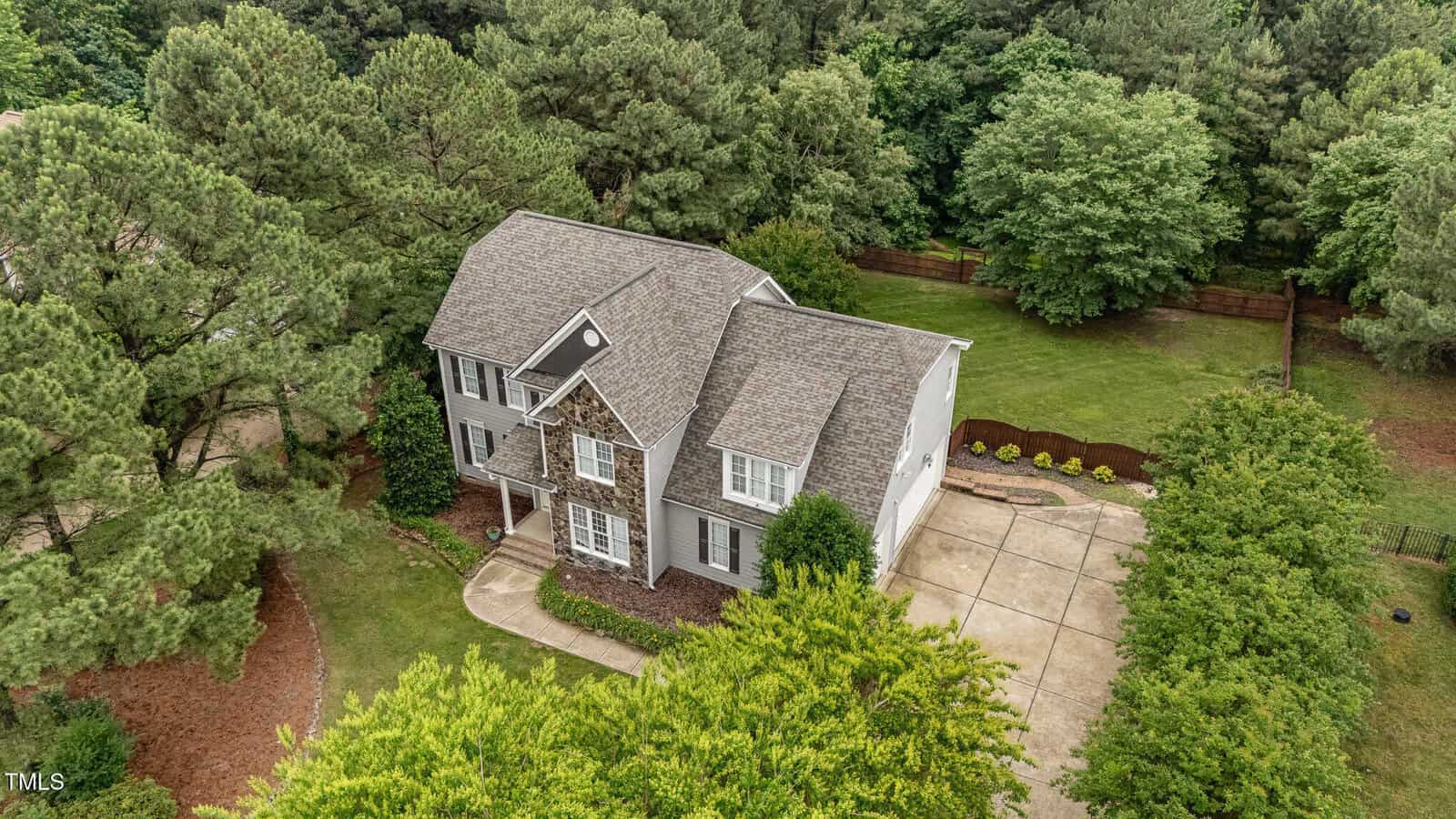 Aerial view of a two-story house with gray siding, stone accents, and a gable roof, surrounded by lush green trees and a spacious driveway. The backyard is grassy and bordered by a wooden fence.