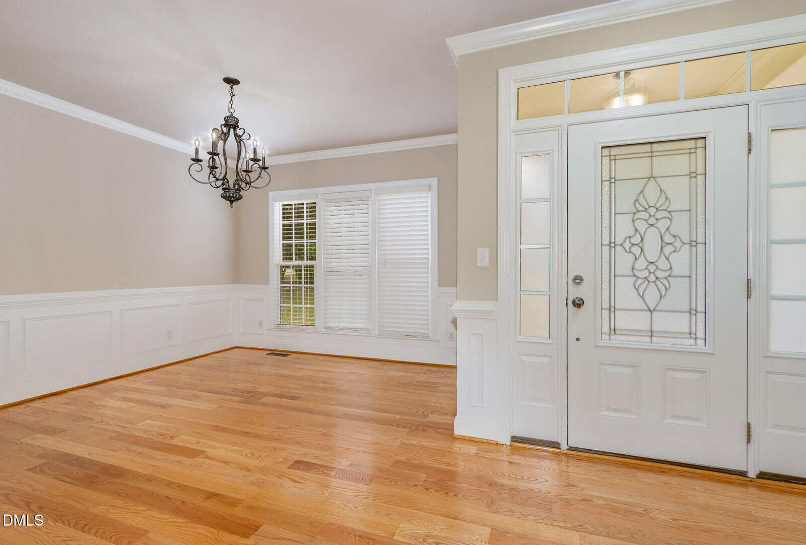 A bright, empty room with light wood floors, beige walls, white wainscoting, a decorative chandelier, and a white front door with glass panels and sidelights. Three large windows with white shutters let in natural light.